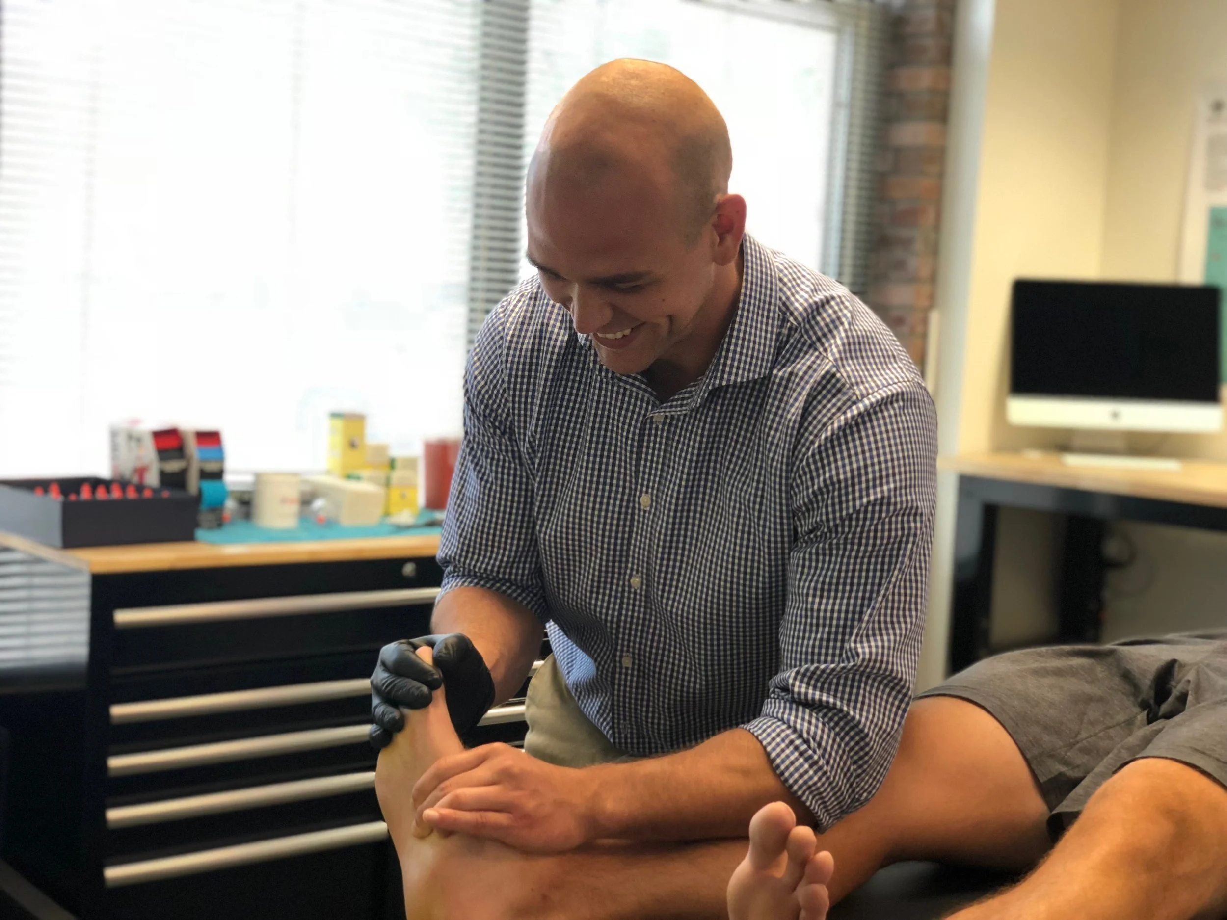 A man with a shaved head wearing a checkered shirt smiling while performing a tattoo on a person's arm in a well-lit room with office equipment in the background. Jon denning