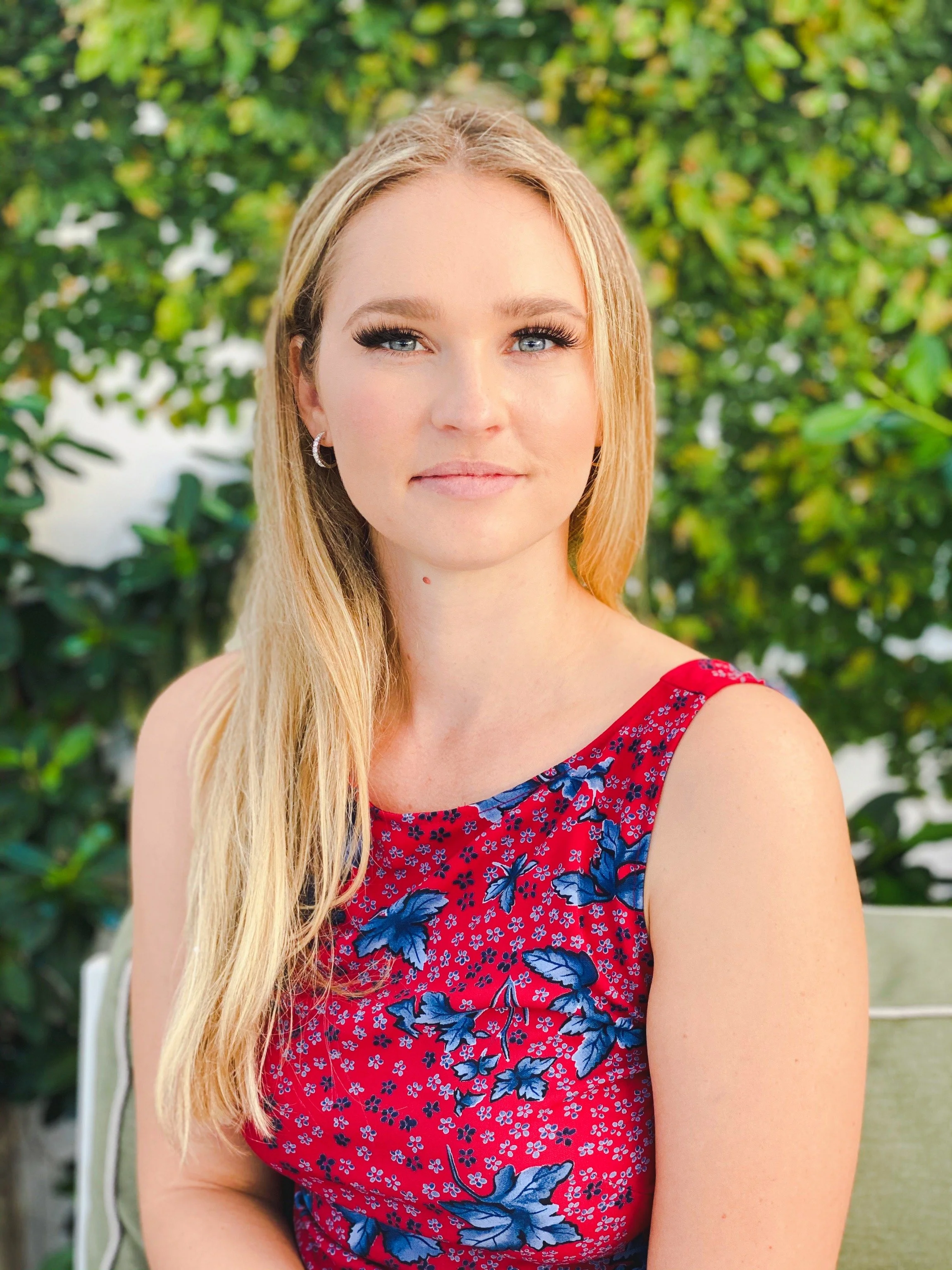 A young woman with long blonde hair and blue eyes, wearing a red sleeveless floral top, standing outdoors with greenery in the background.