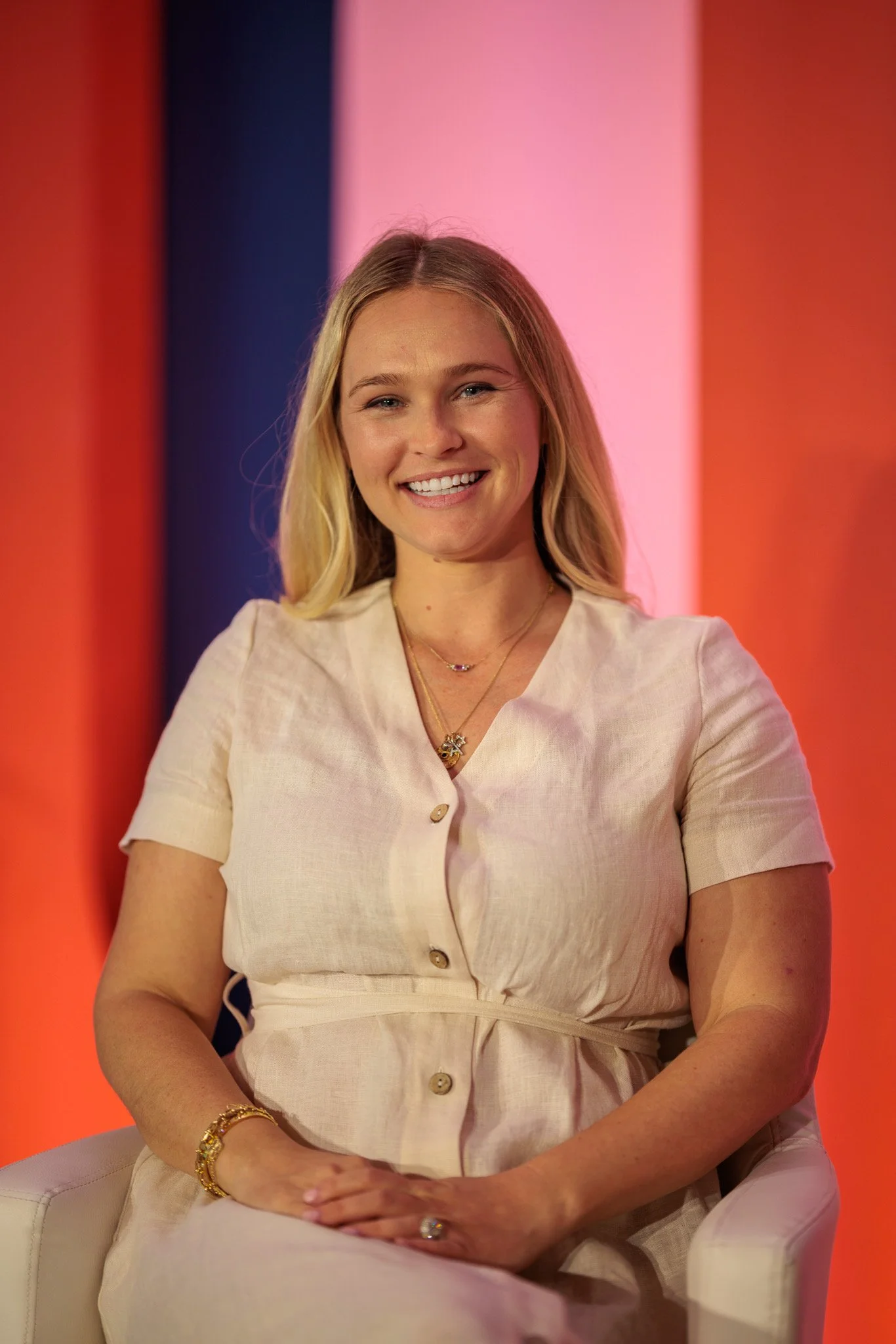 A young woman with long blonde hair, smiling, sitting on a white chair against a colorful background with red, black, pink, and purple accent colors.