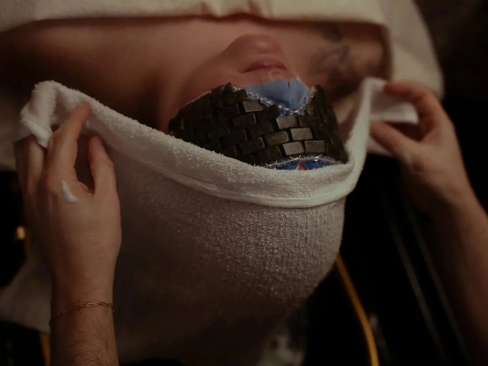 A person lying down with their face partially covered by a cloth, a puzzle piece is being placed over their mouth during a rope challenge or game.