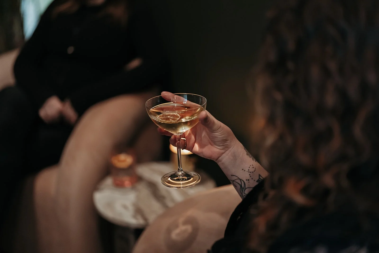 Person holding a champagne glass with a slice of lemon in it, with other people sitting nearby in a dimly lit setting.