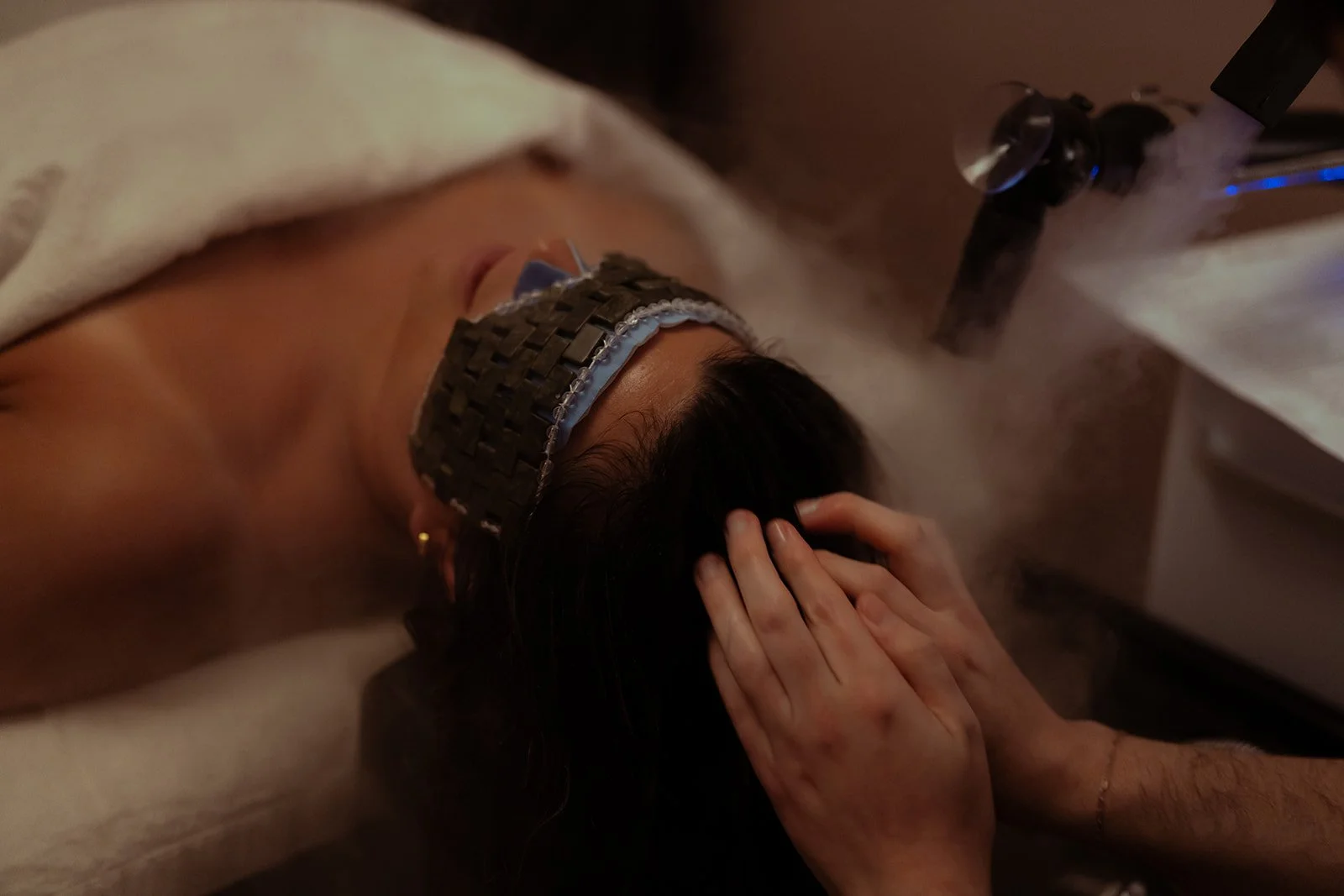 A person lying face down on a table receives a steam facial treatment, with a facial mask and a steam device emitting vapor.