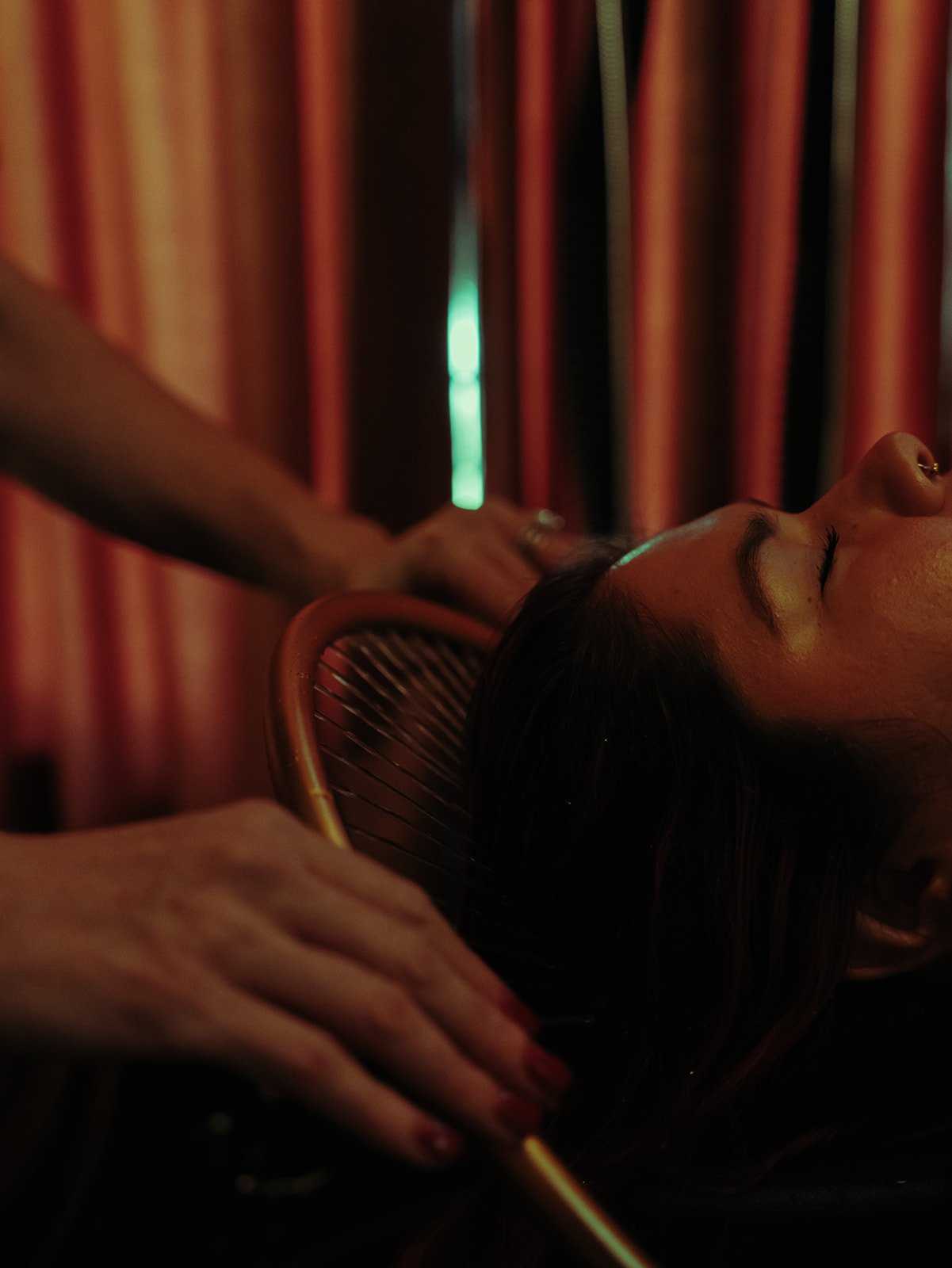 A woman receives a massage while lying on a chair with her eyes closed, in a dimly lit room with red curtains.
