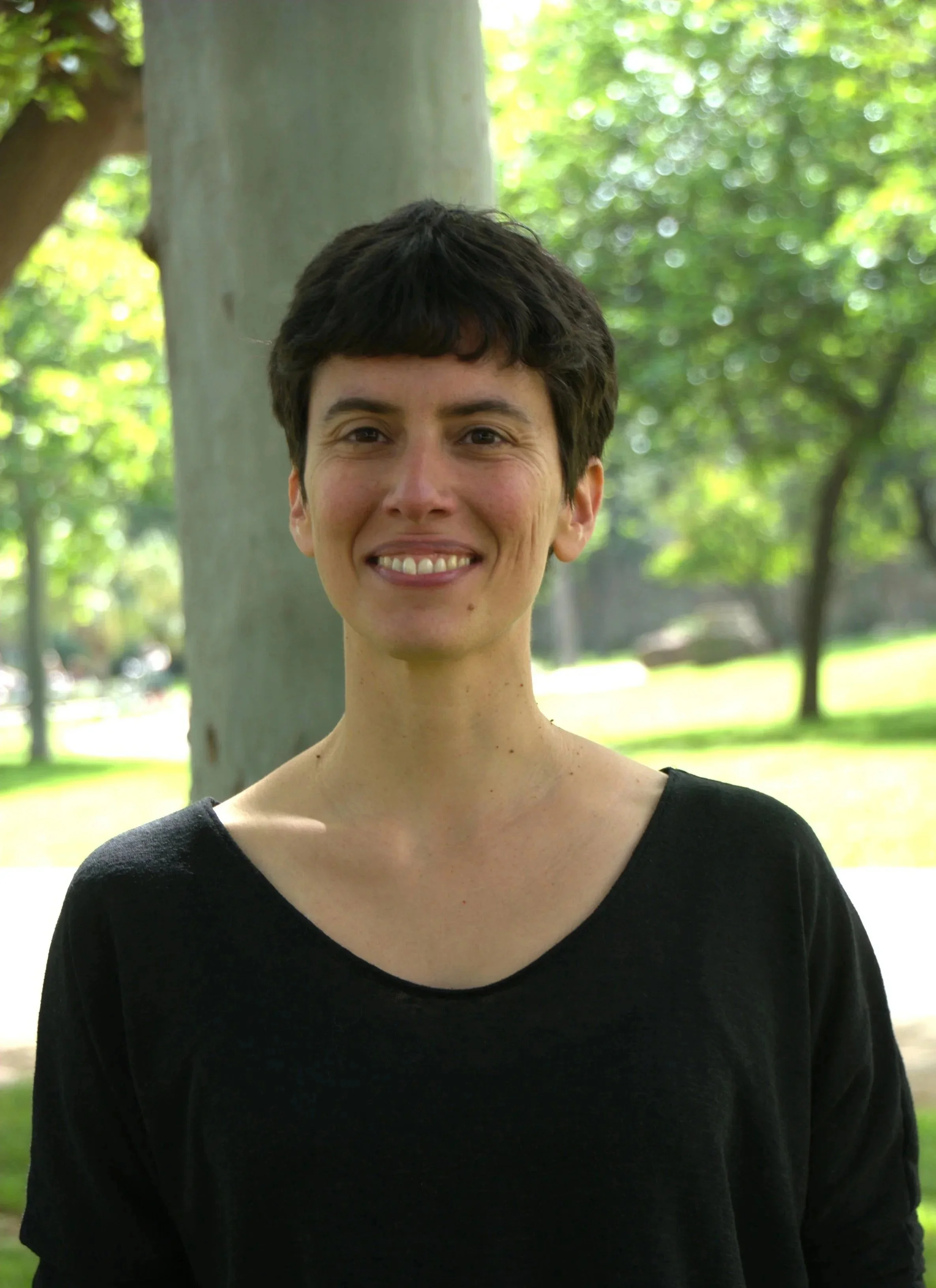 A woman with short dark hair smiling outdoors in a park with green trees in the background.