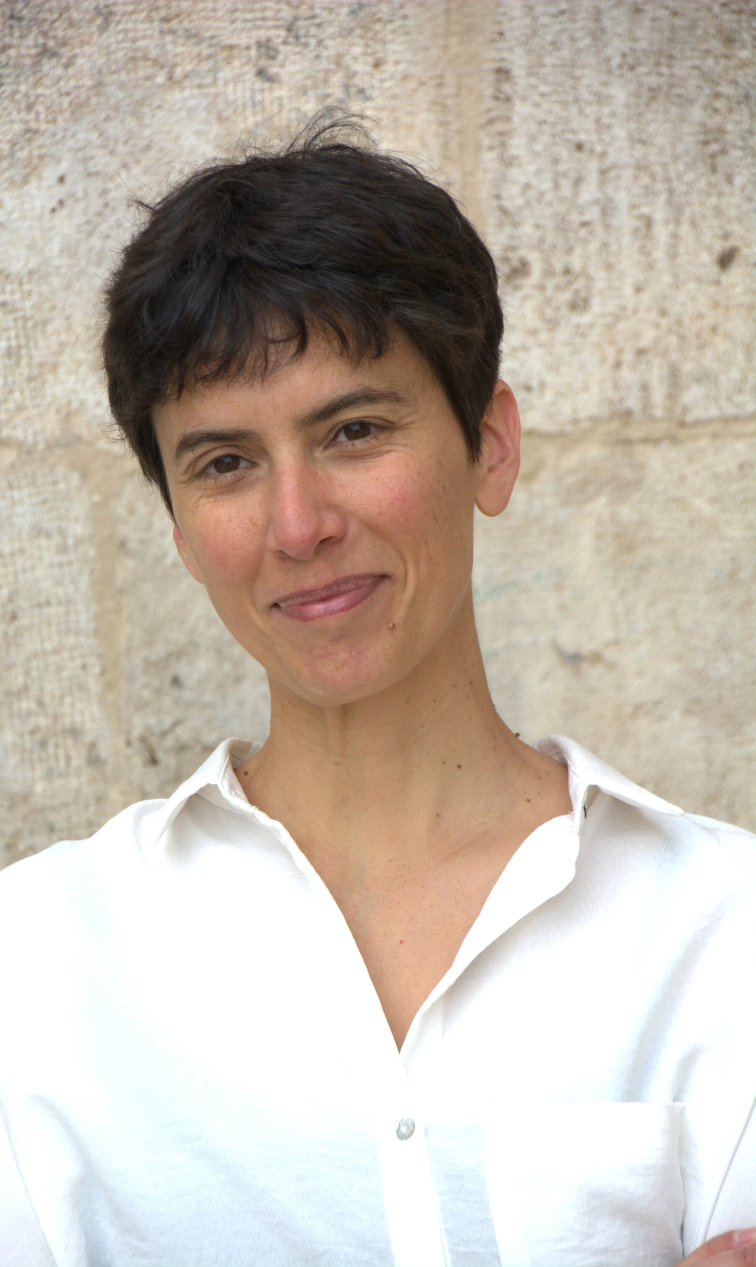 A woman with short dark hair wearing a white shirt standing outdoors in front of a stone wall.