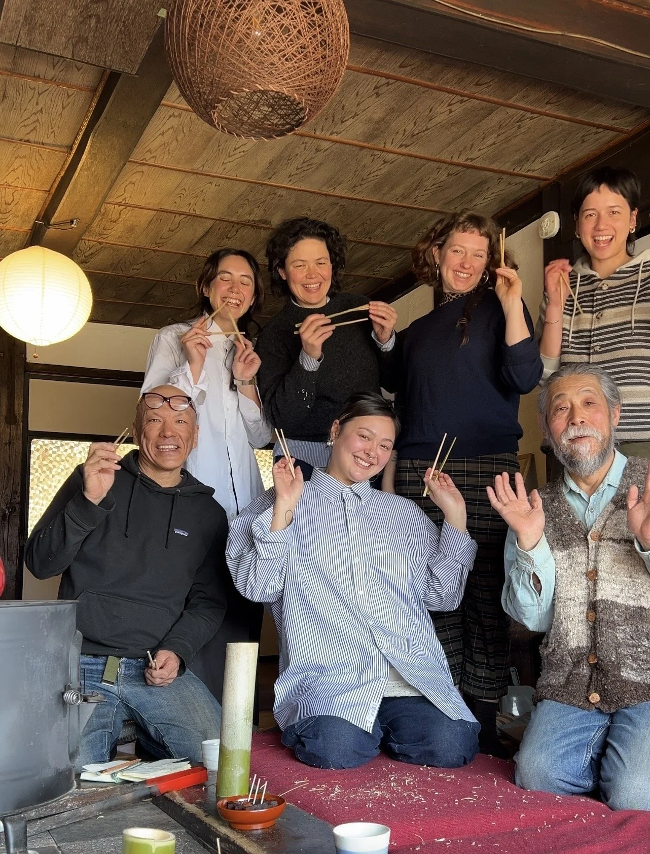 A group of nine people gathered together in a restaurant, smiling and holding chopsticks. They are sitting and standing in front of a wooden ceiling with hanging lights. The table in front has some chopsticks, a small bowl, and a green drink.