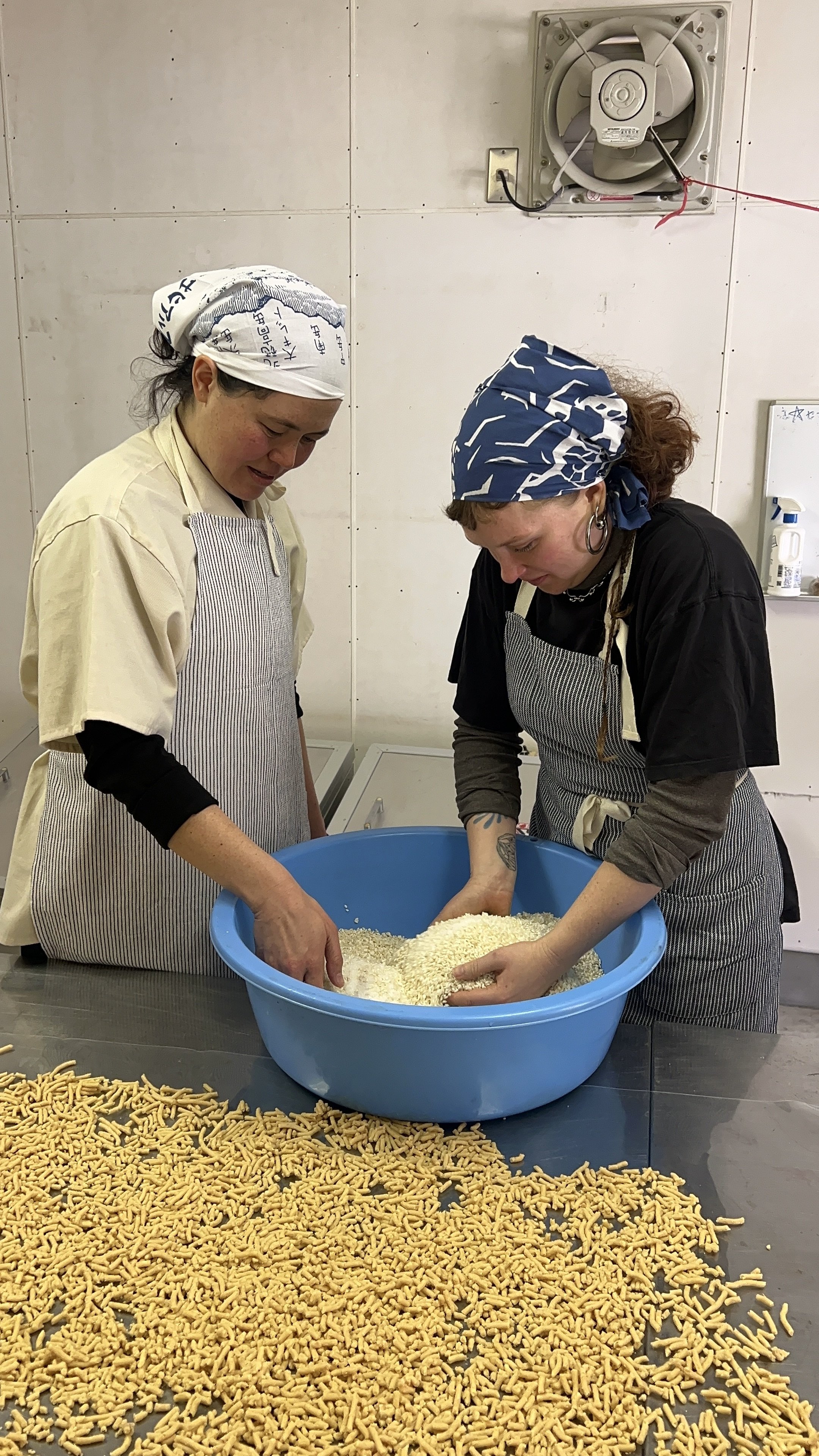 Two women wearing aprons and headscarves preparing a large quantity of pasta or rice in a blue tub at a kitchen or food production facility, with pasta or breadsticks scattered on the table.