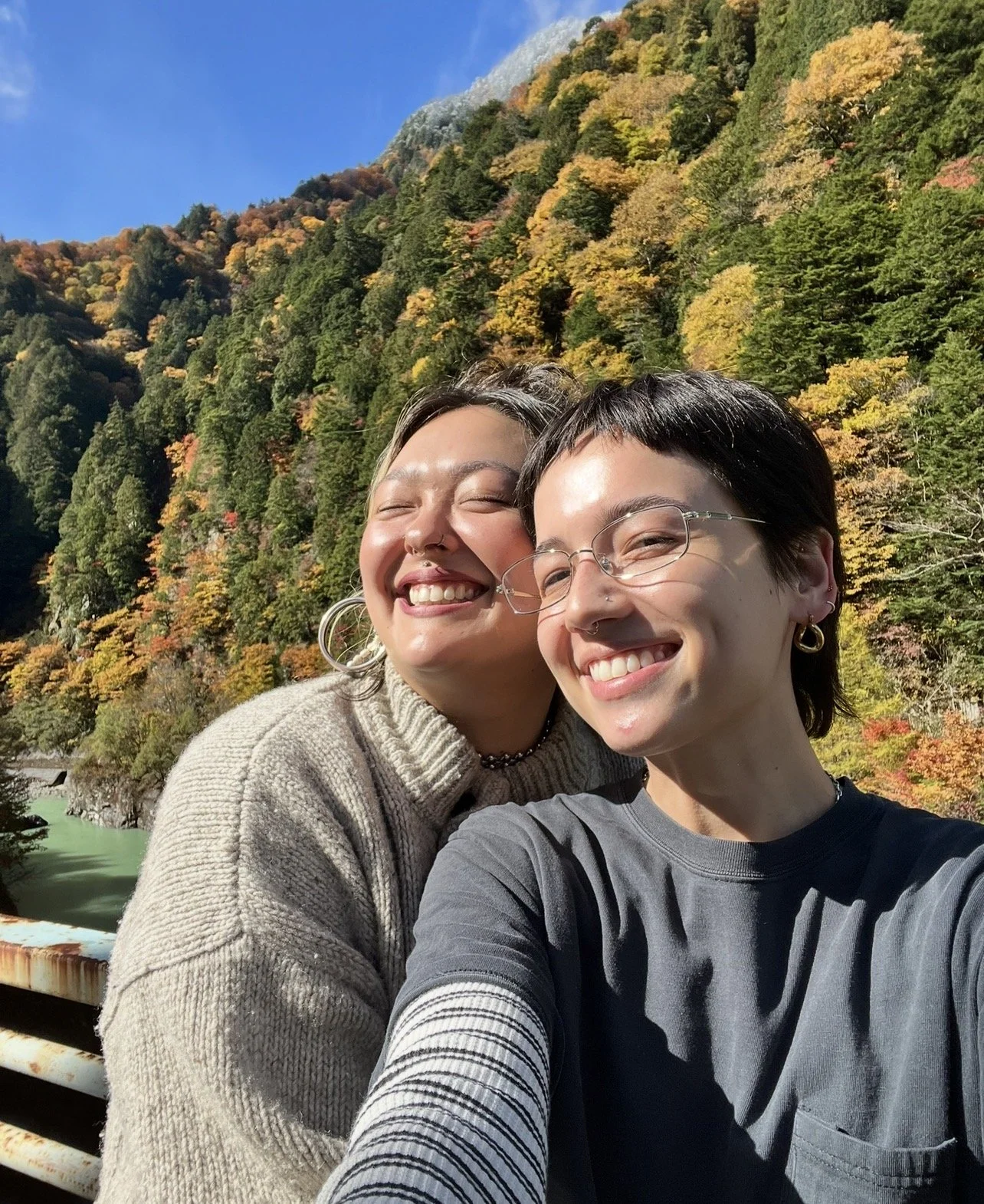 Two smiling women taking a selfie outdoors with a colorful autumn forest and mountain in the background.