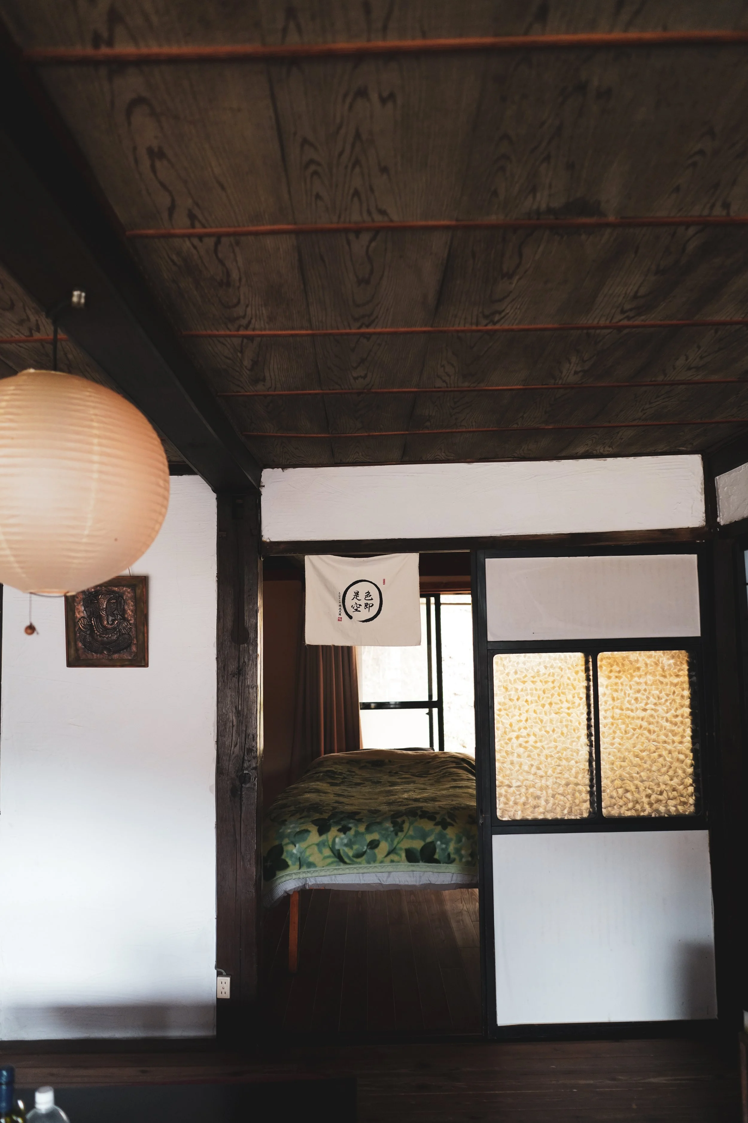 Interior of a room with a dark wooden ceiling, a hanging paper lampshade on the left, and a sliding door with a frosted glass panel that has a decorative pattern. There is a bed with a green floral bedspread partially visible, and a window with brown