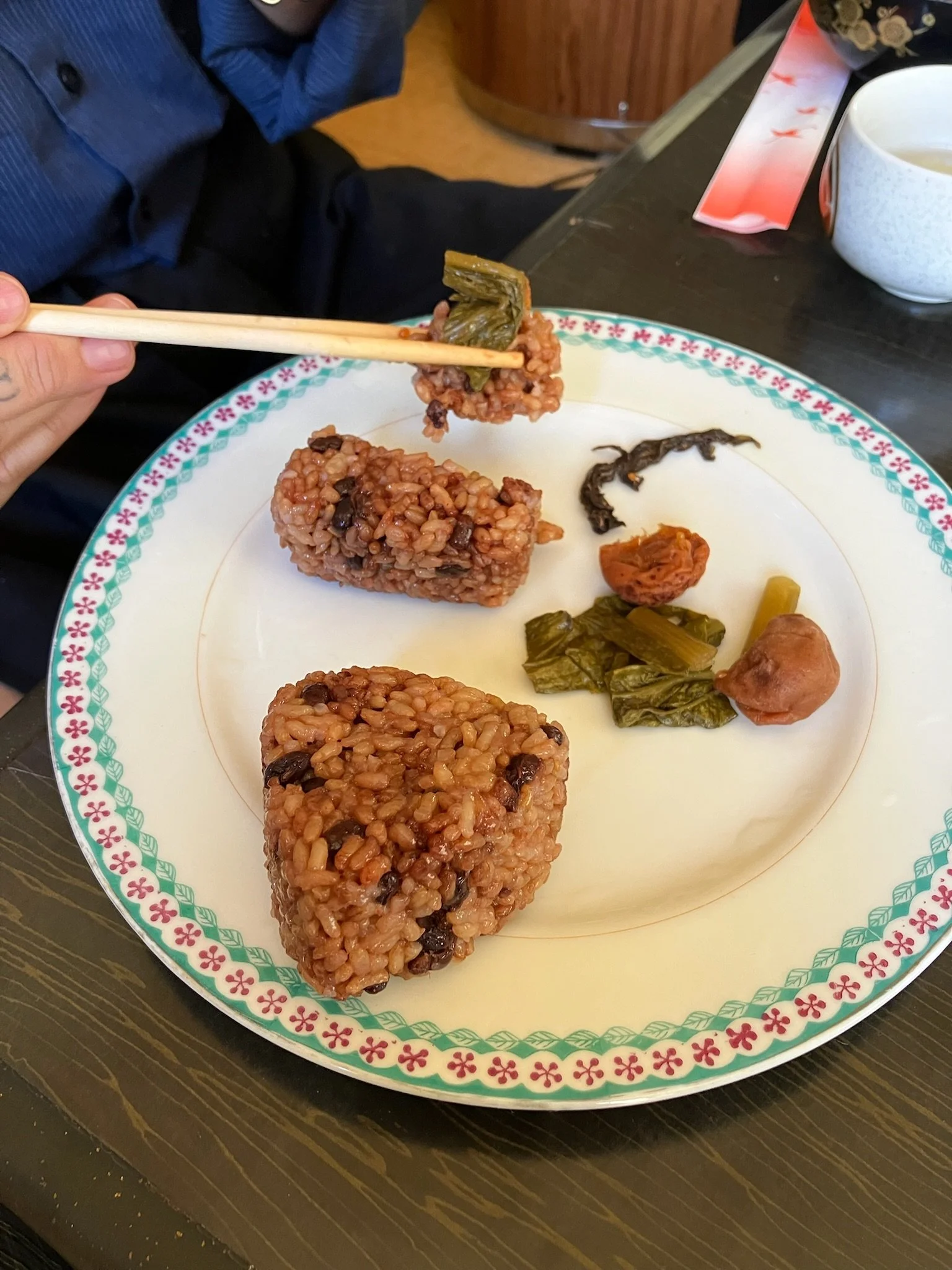 Plate with three rectangular rice cakes and a few pickled vegetables, with a person using chopsticks to hold one rice cake.