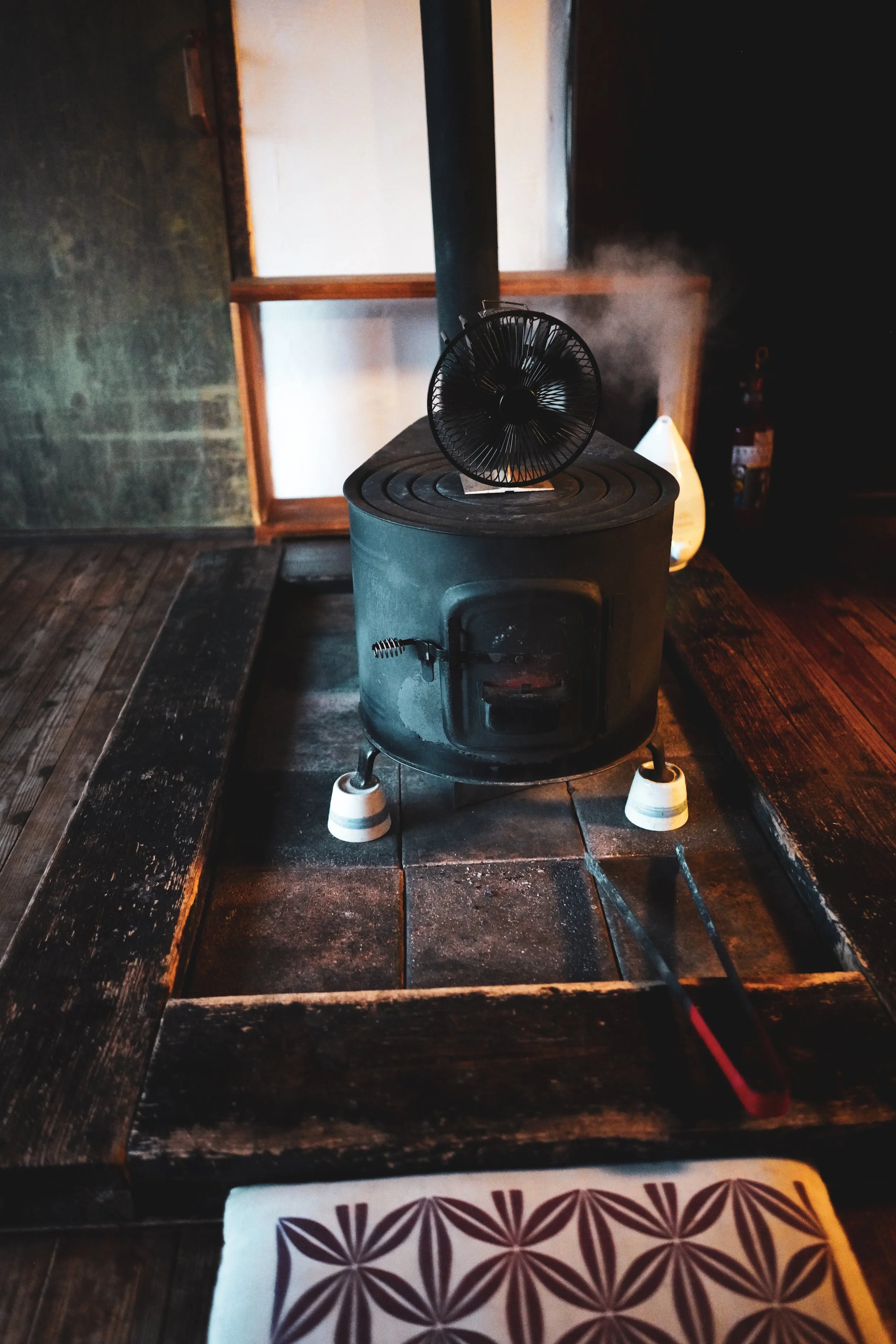 A small black wood stove with a vent pipe emitting steam or smoke, placed on a wooden floor in a rustic room, with a decorative cushion in the foreground.