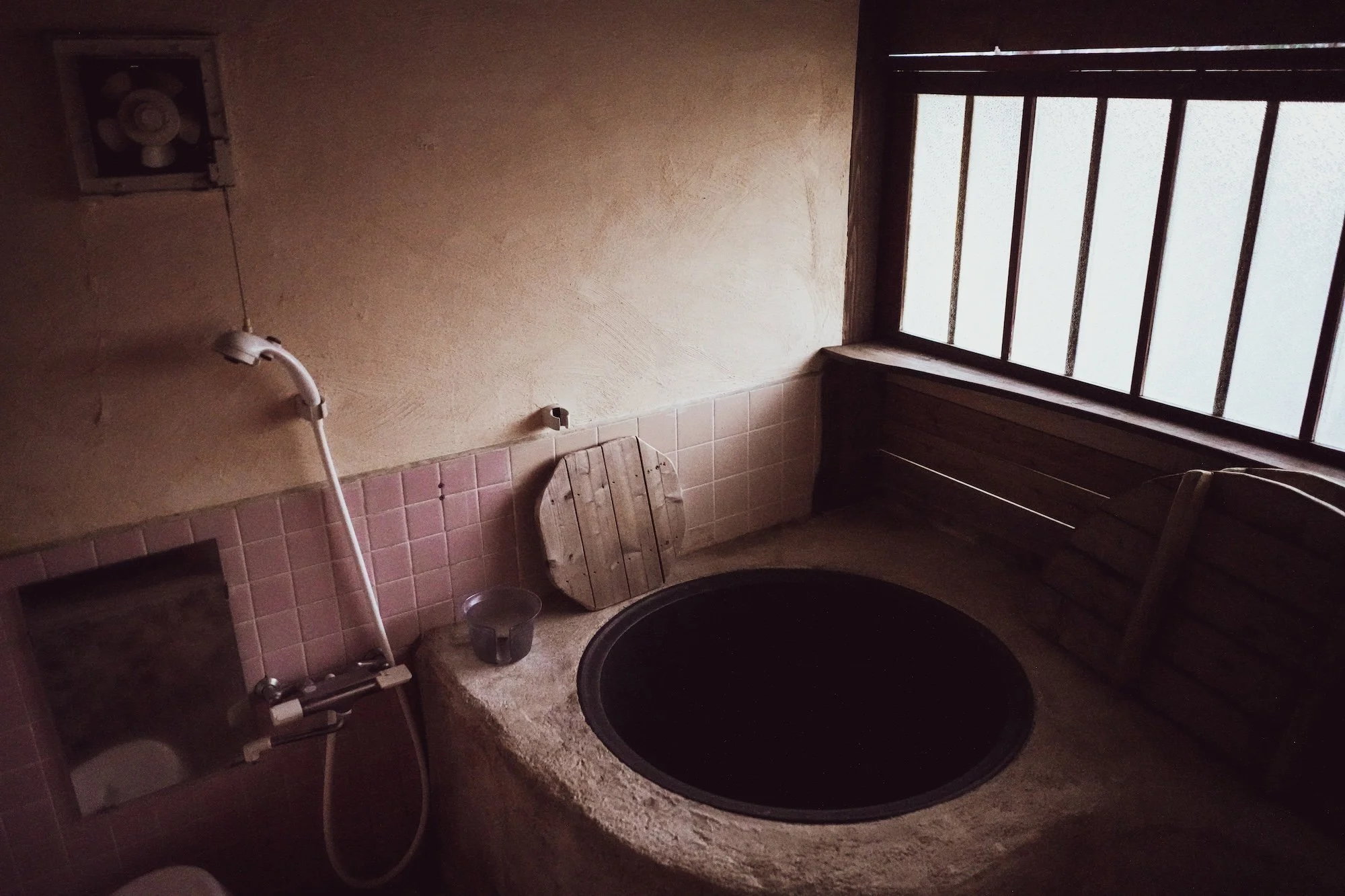 Bathroom with pink tiled lower wall and beige upper wall, featuring a squat toilet with a black seat, a wooden stool, a bucket, a small stool, and a window with bars allowing light into the room.