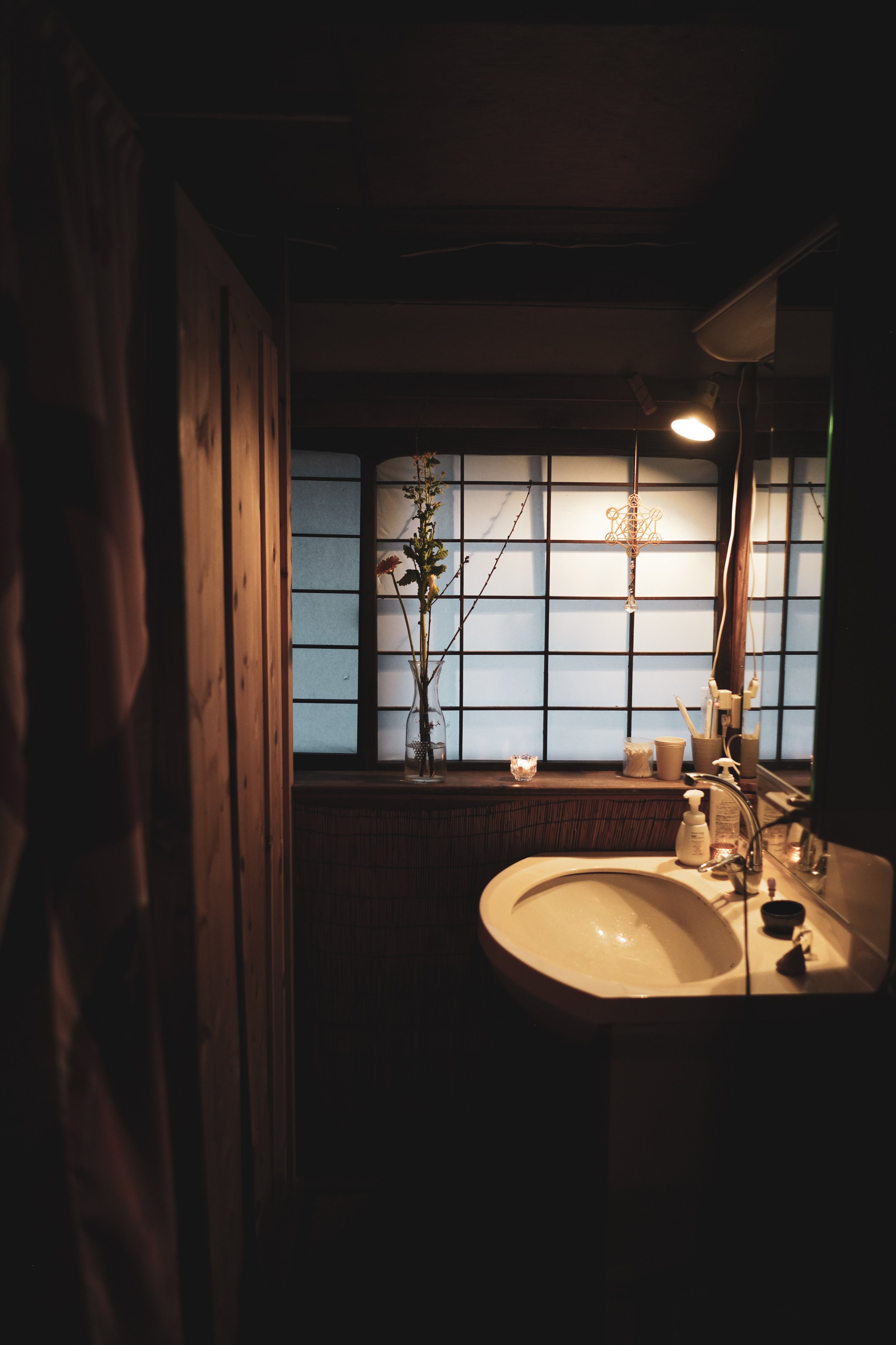 A bathroom with a sink, soap dispenser, toothbrush holder, and small decorative items on the counter. Behind the sink is a window with frosted glass and a hanging cross-shaped ornament. A vase with flowers sits on the ledge beneath the window. The ro