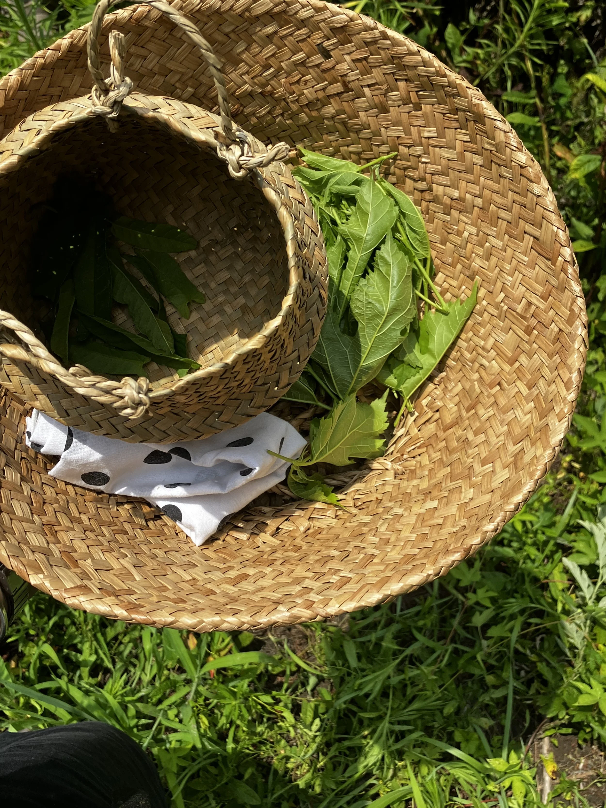 A wicker hat and a woven basket filled with green leaves on grass.