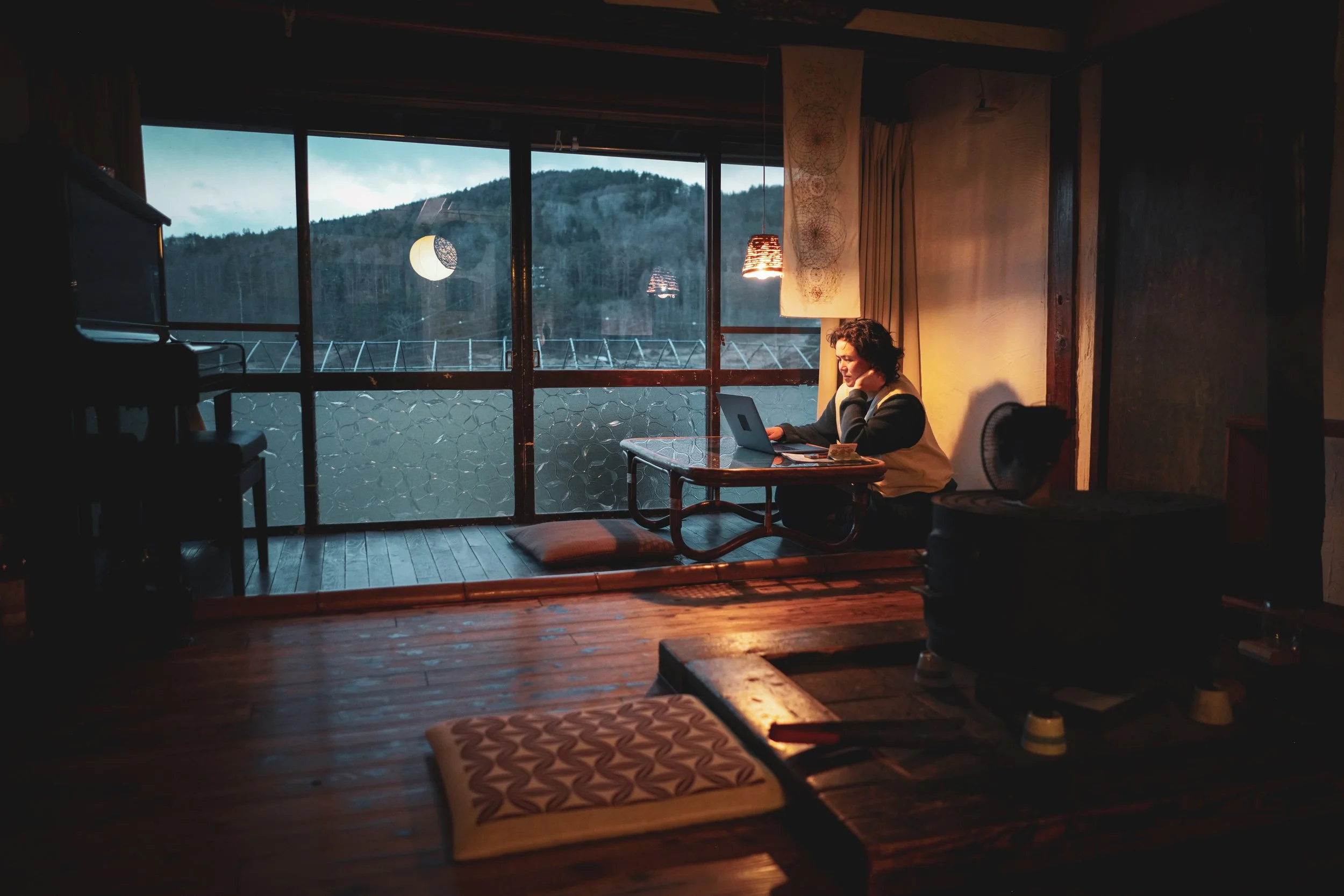 A person sitting at a table working on a laptop in a cozy room with a large window showing a scenic view of a mountain and trees. The room is dimly lit with hanging lamps and some decorative items on the walls.