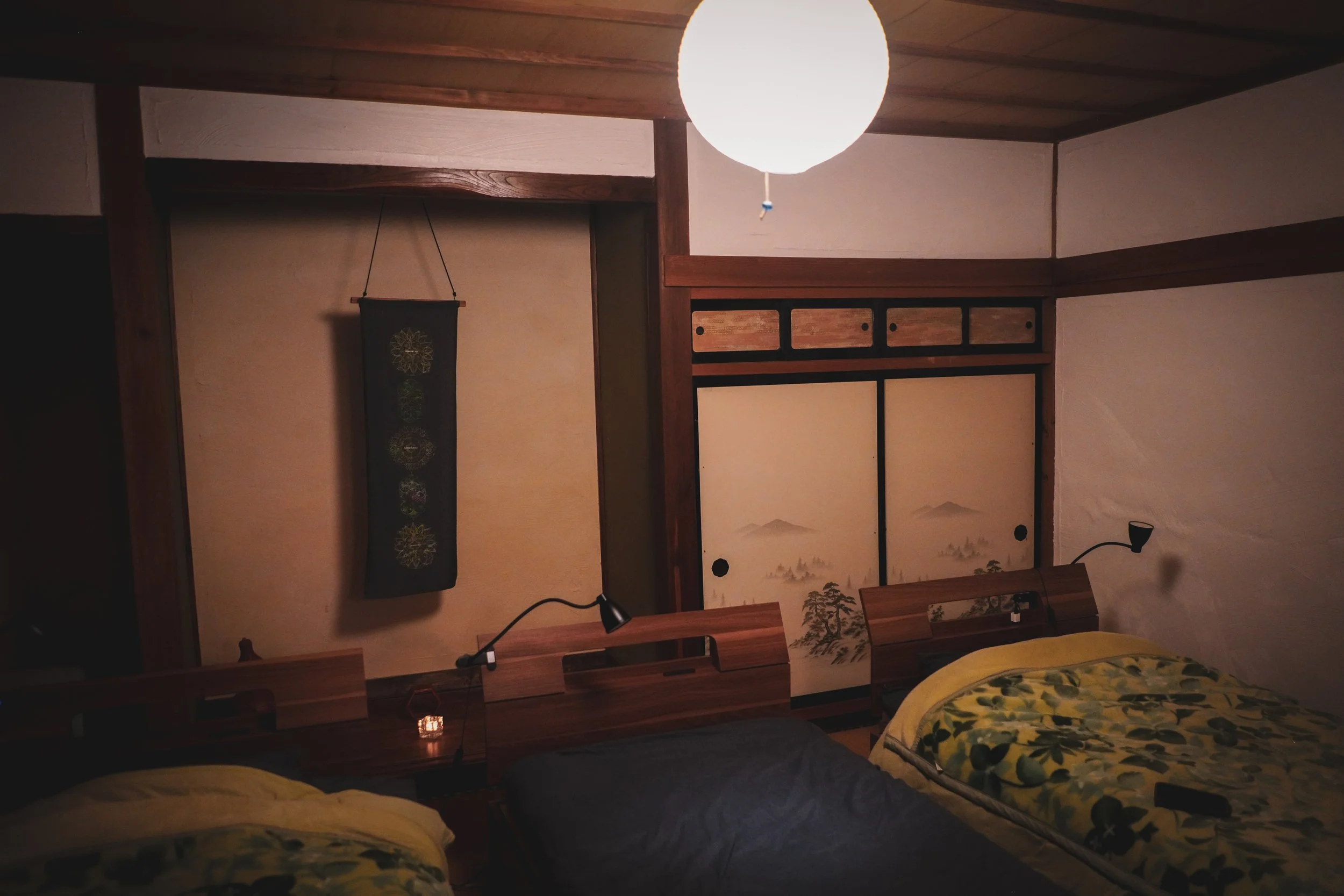 A bedroom with wooden frames and traditional Japanese sliding doors, a large round ceiling light, two beds with yellow and dark gray bedding, a small hanging scroll on the wall, and a small lamp on the bedside table.