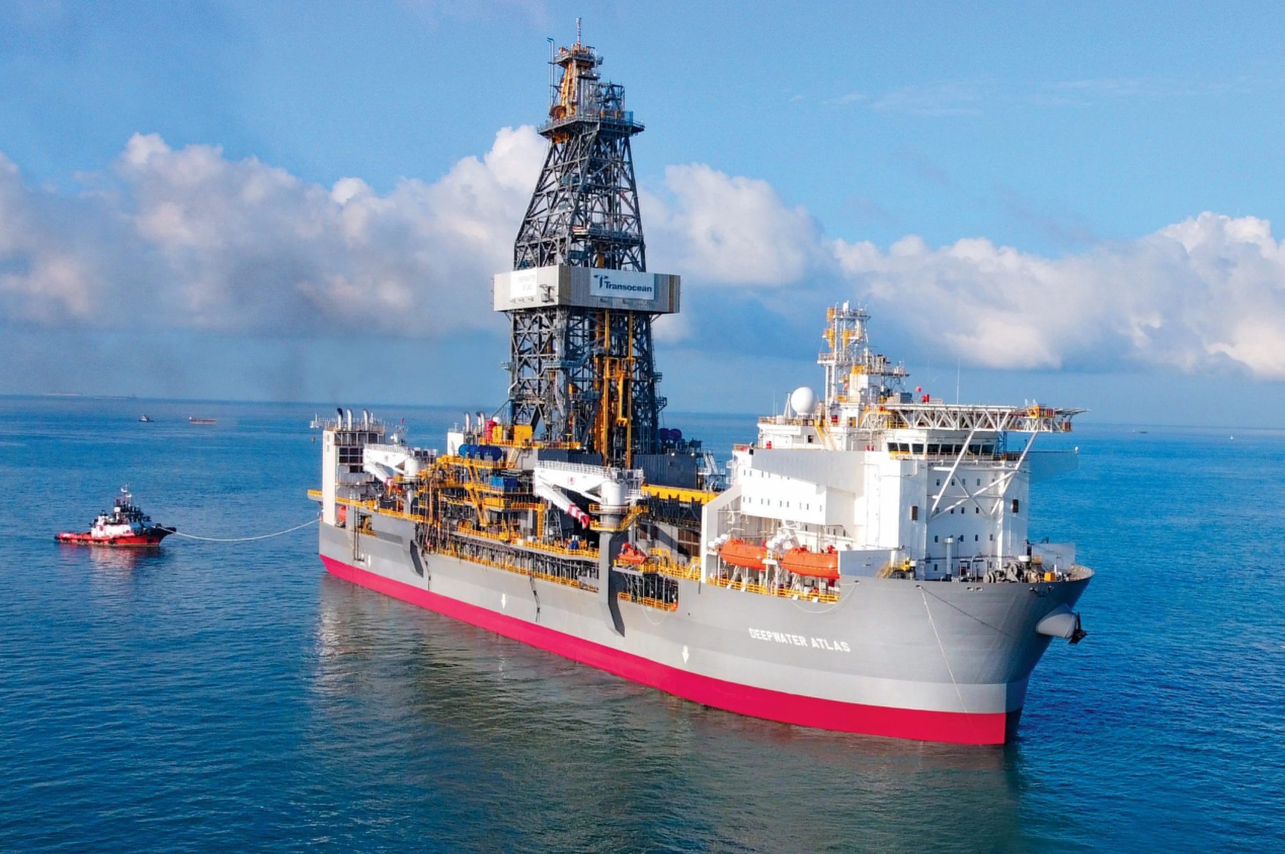 Offshore oil drilling rig named Deepwater Atlas with a support vessel attached, floating on calm ocean water with a partly cloudy blue sky in the background.