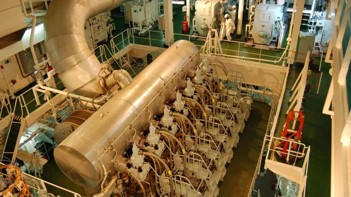 Large industrial machinery on a ship's engine room with pipes, valves, and crew members in protective gear.
