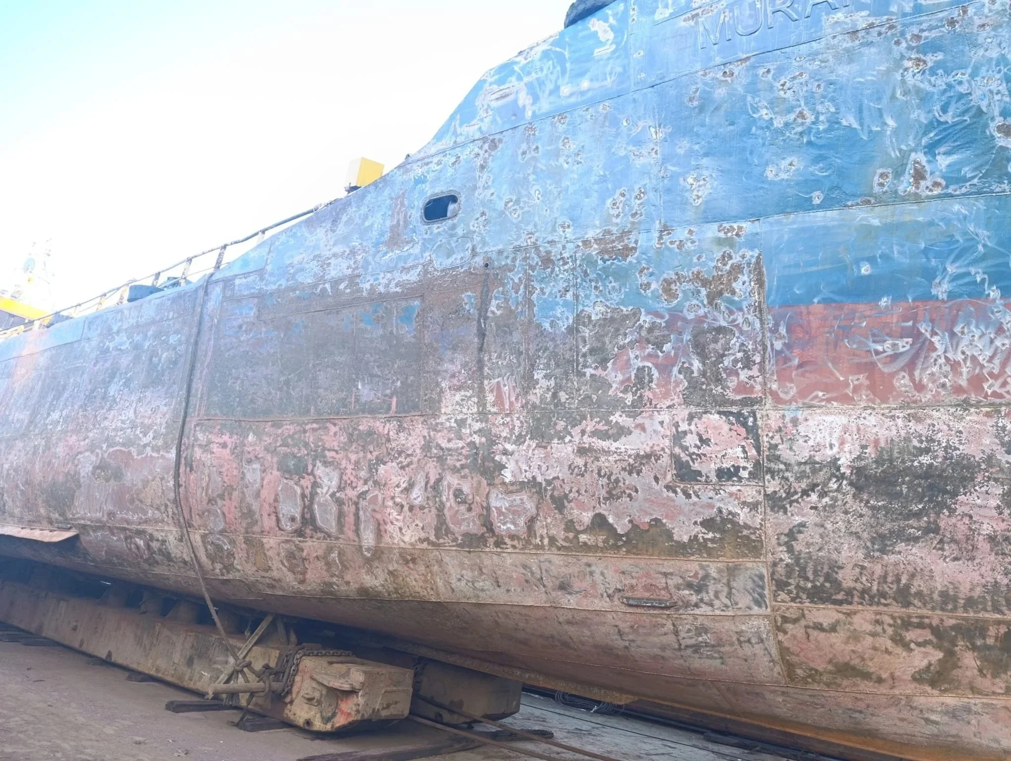 Close-up of an old, weathered boat hull with peeling paint and corrosion, sitting on wooden supports.