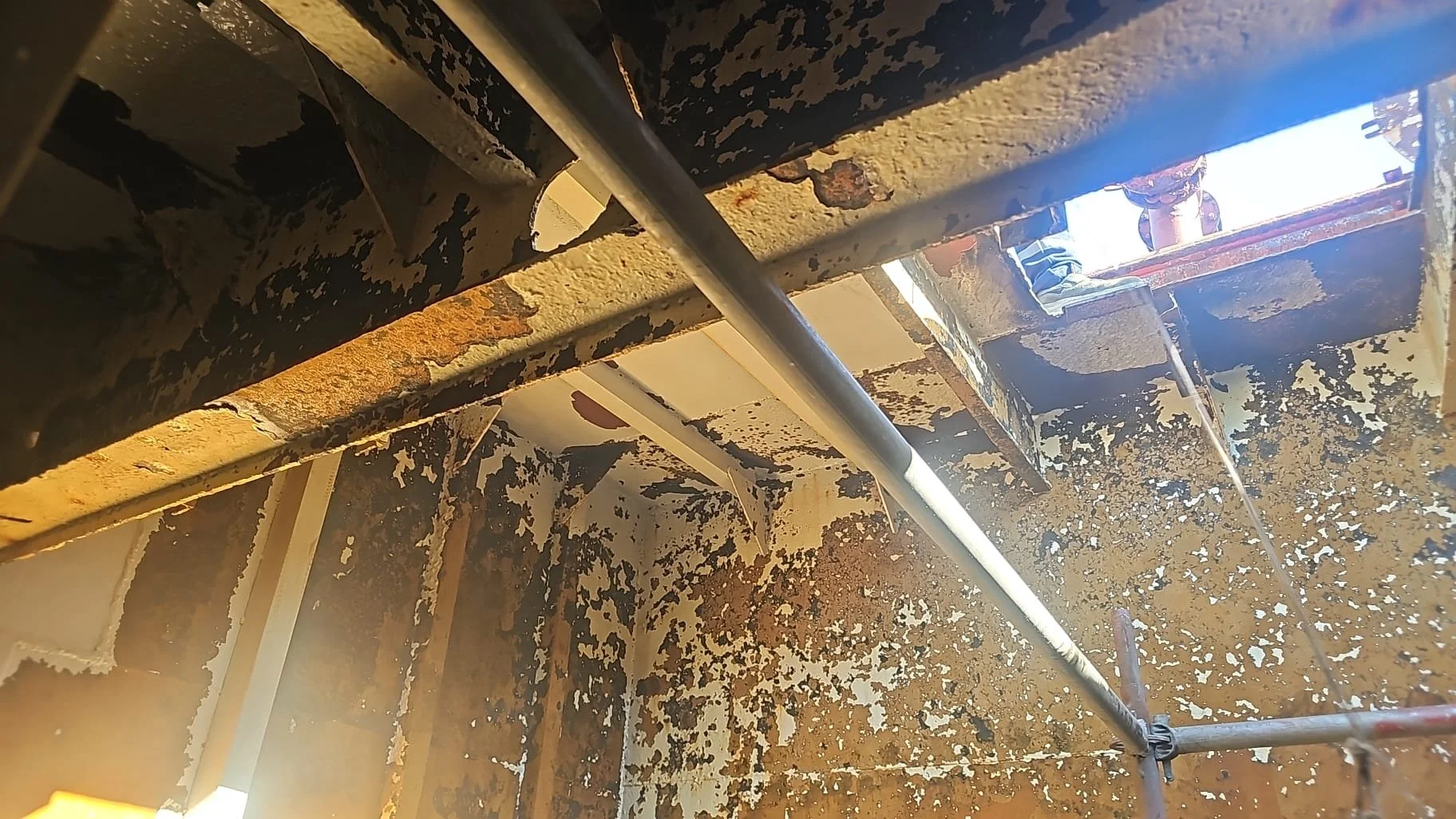 A rusted and peeling interior wall with scaffolding and an opening at the top allowing sunlight to enter.