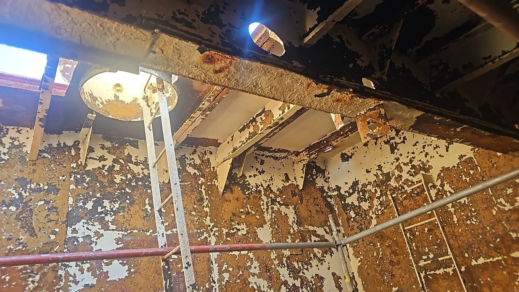A rusted, deteriorated interior ceiling with peeling paint, ladders, and scaffolding, showing corrosion and decay.