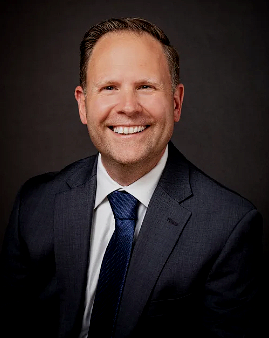 Headshot of a smiling man in a dark suit, white shirt, and blue tie against a dark background.