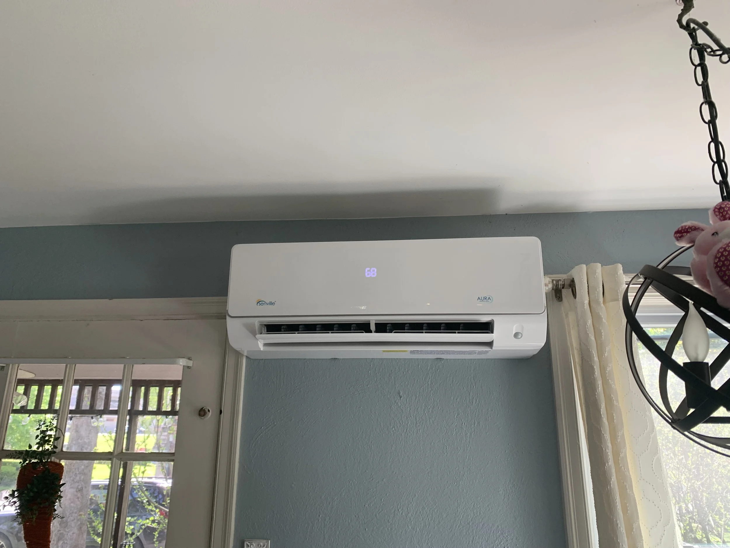 Wall-mounted white air conditioner unit with digital display reading 68, situated above a window with beige curtains, in a room with teal walls and a nearby chandelier with pink plush flowers.