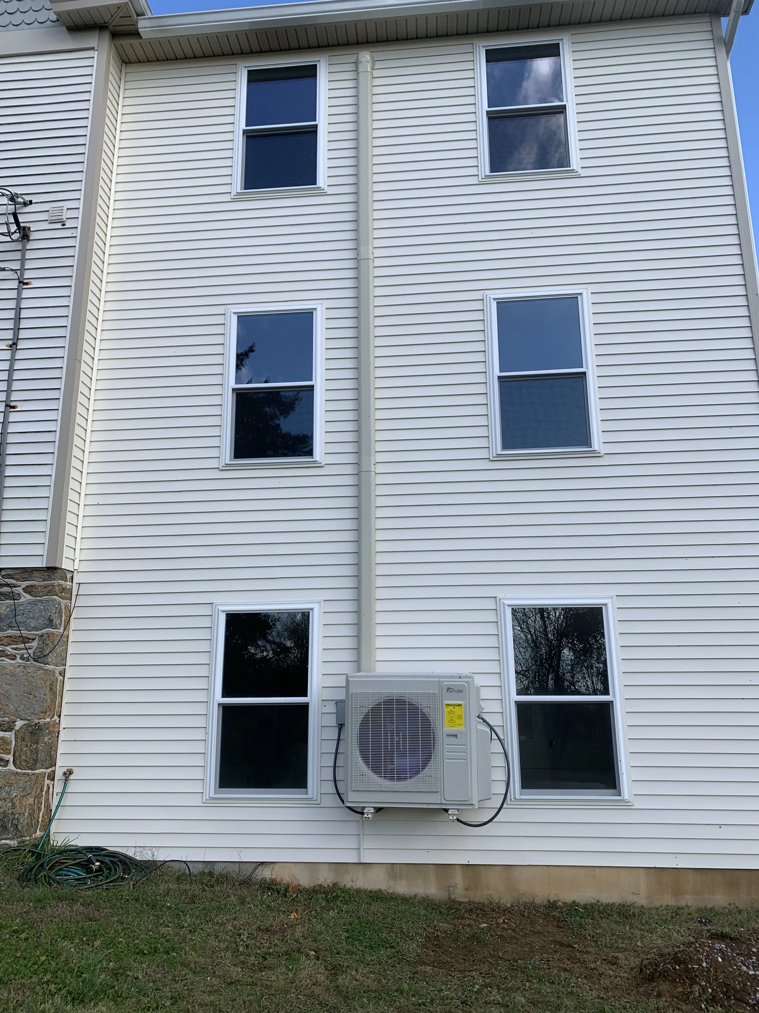 Exterior of a multi-story house with white vinyl siding, six windows, and an air conditioning unit mounted on the wall.