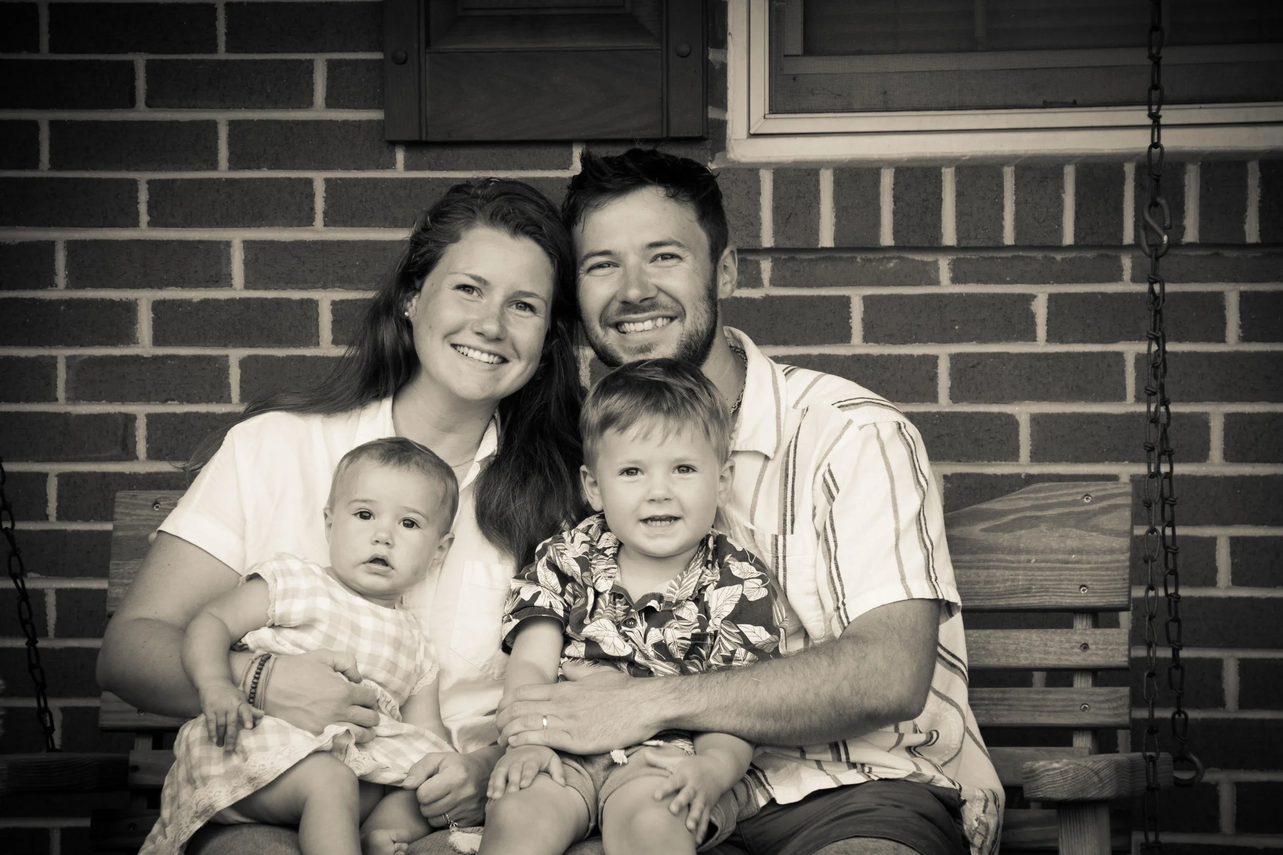 A family of four smiling and sitting on a wooden swing in front of a brick wall, with two young children and two adults.