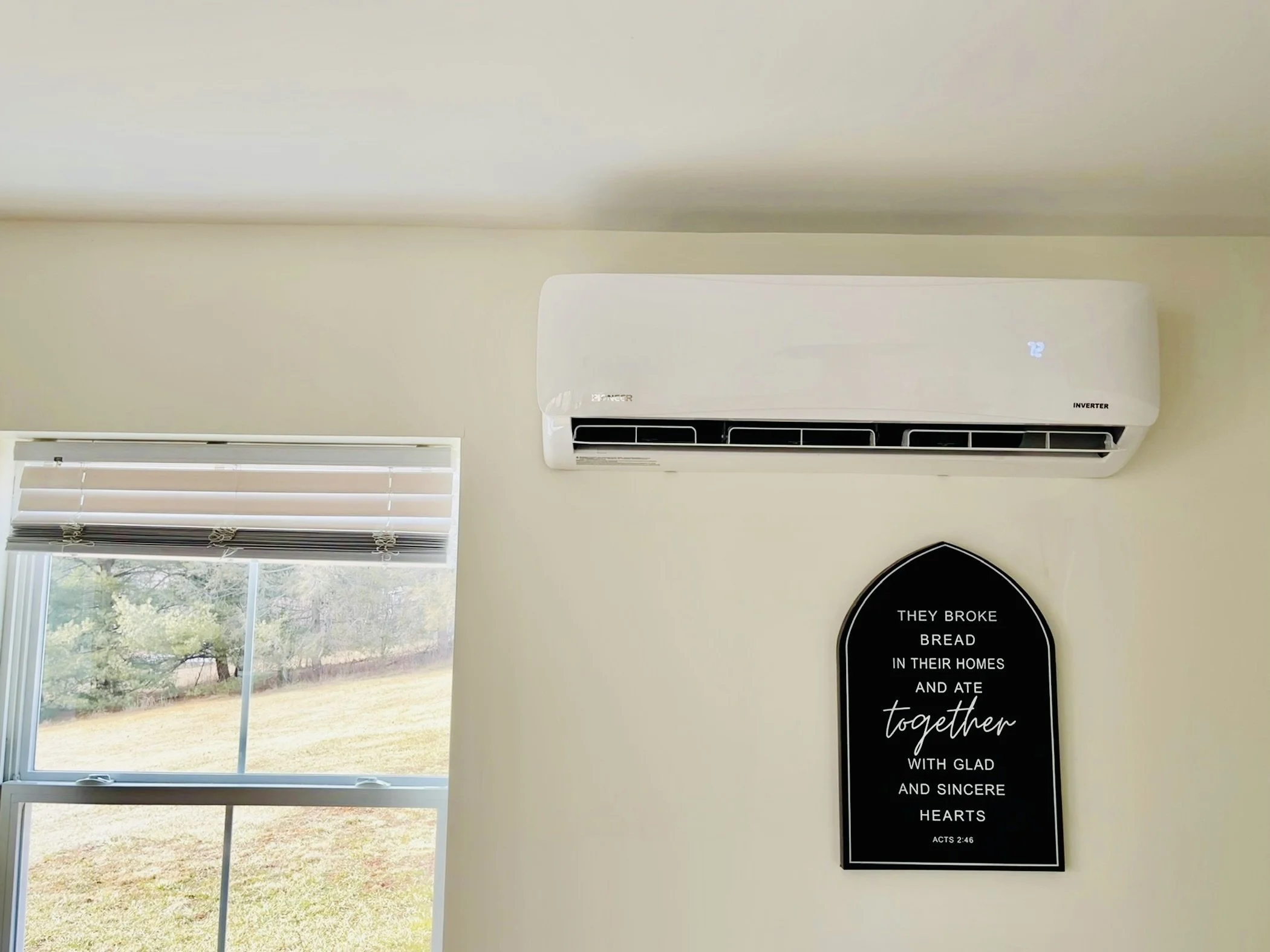 A white wall-mounted air conditioner unit with digital display, a window with blinds, and a black plaque with a Bible verse about bread and hearts.