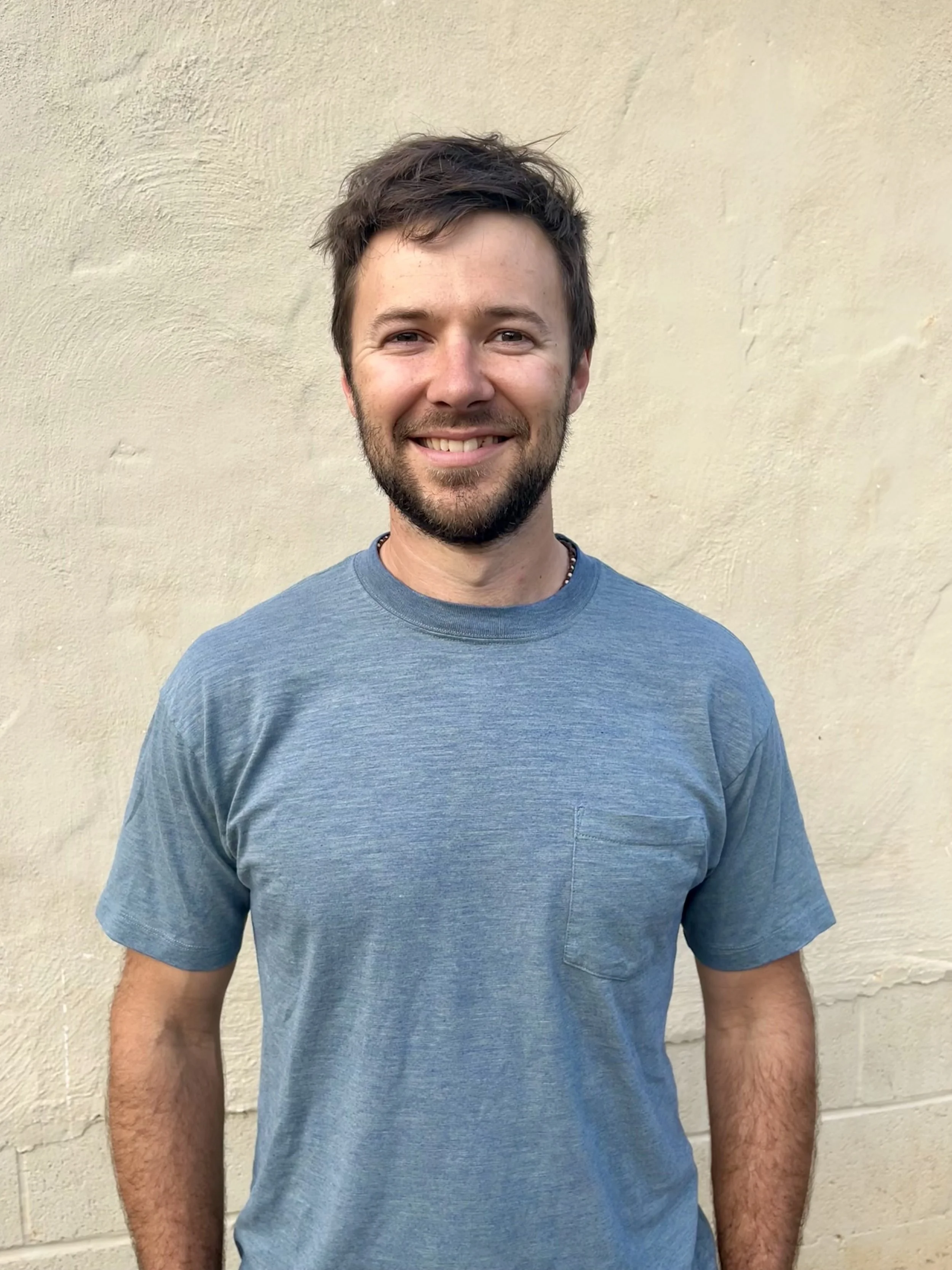A young man with dark hair and a beard smiling, wearing a blue t-shirt, standing against a beige textured wall.