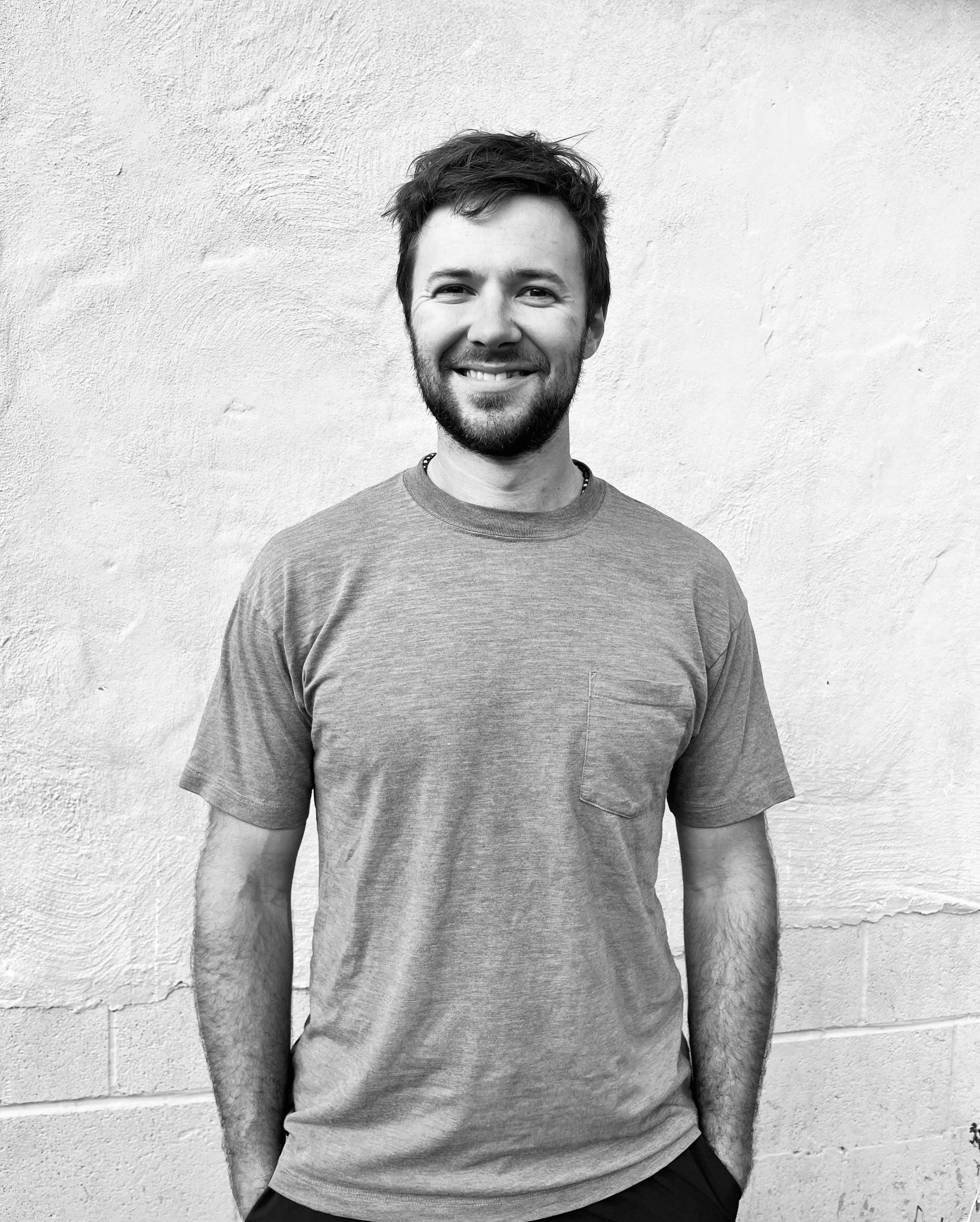 A young man with a beard and tousled hair smiling while standing in front of a textured wall.
