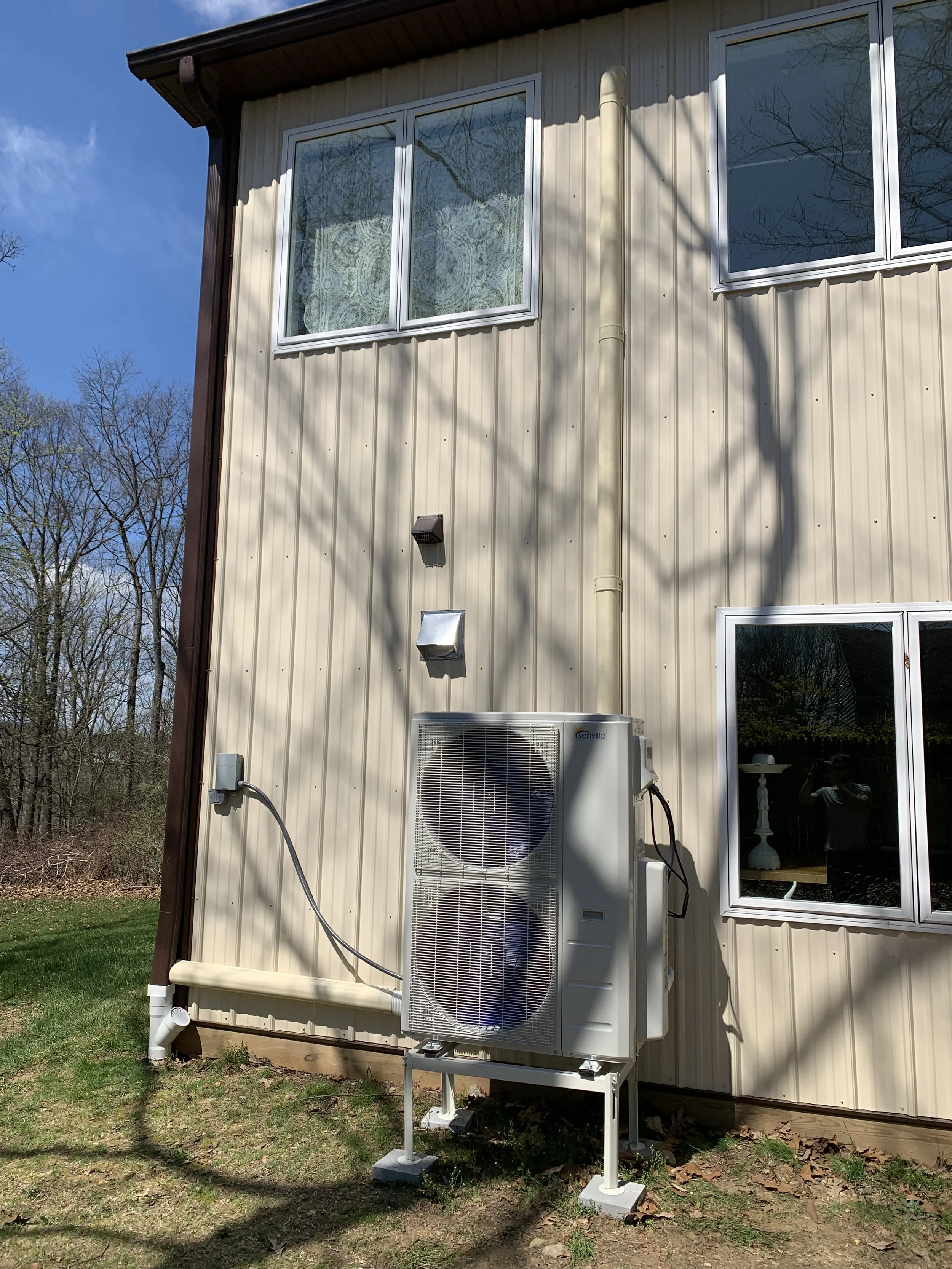 A beige metal-sided house with two windows, one above and one to the right side. An outdoor HVAC unit and a power box are mounted on the wall beneath the windows. Shadows of tree branches are cast on the facade. The area below is grassy with some patches of dirt and fallen leaves.
