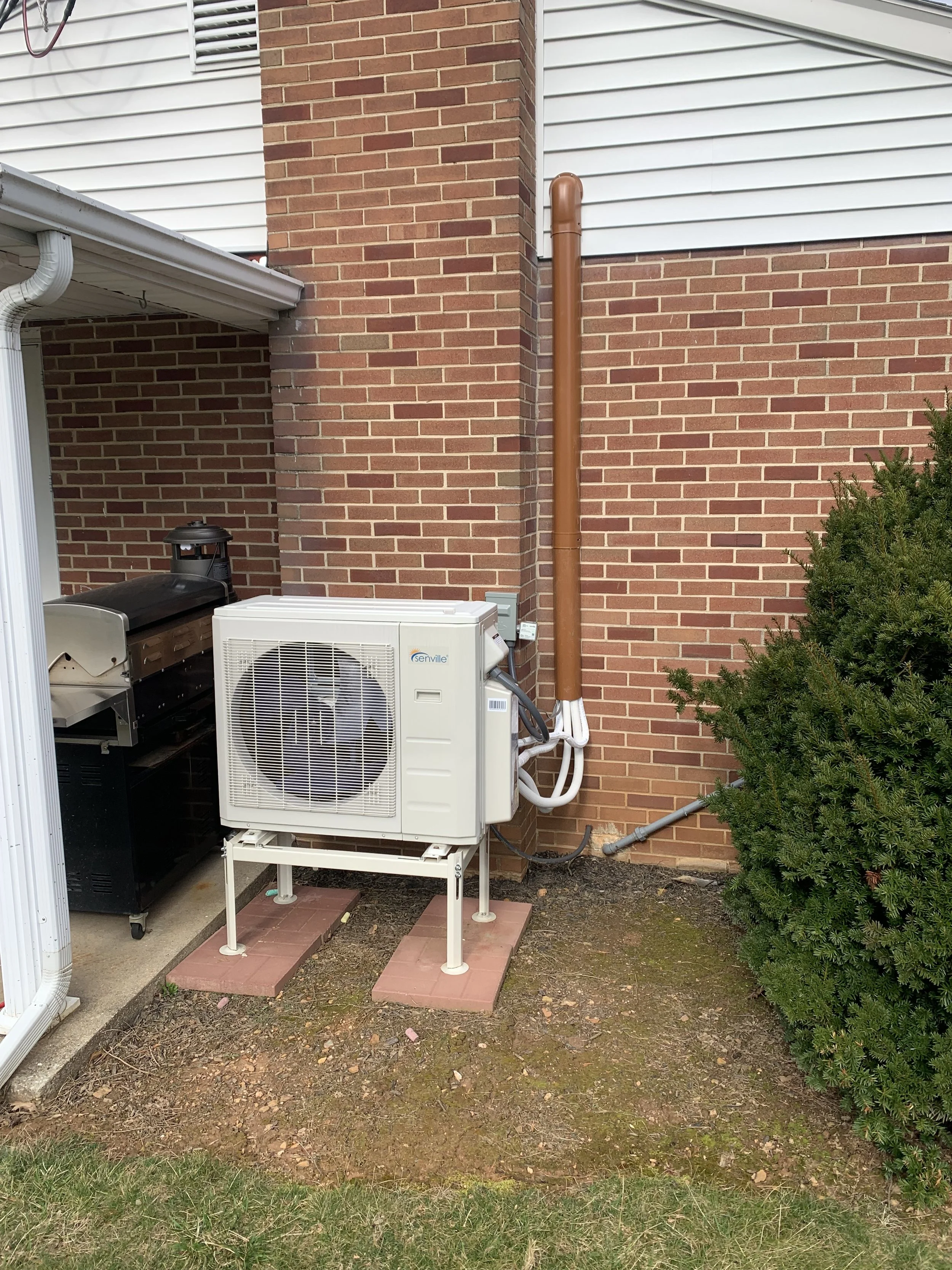Outdoor HVAC unit on elevated bricks with brick and siding house wall, a bush, and some ground dirt.
