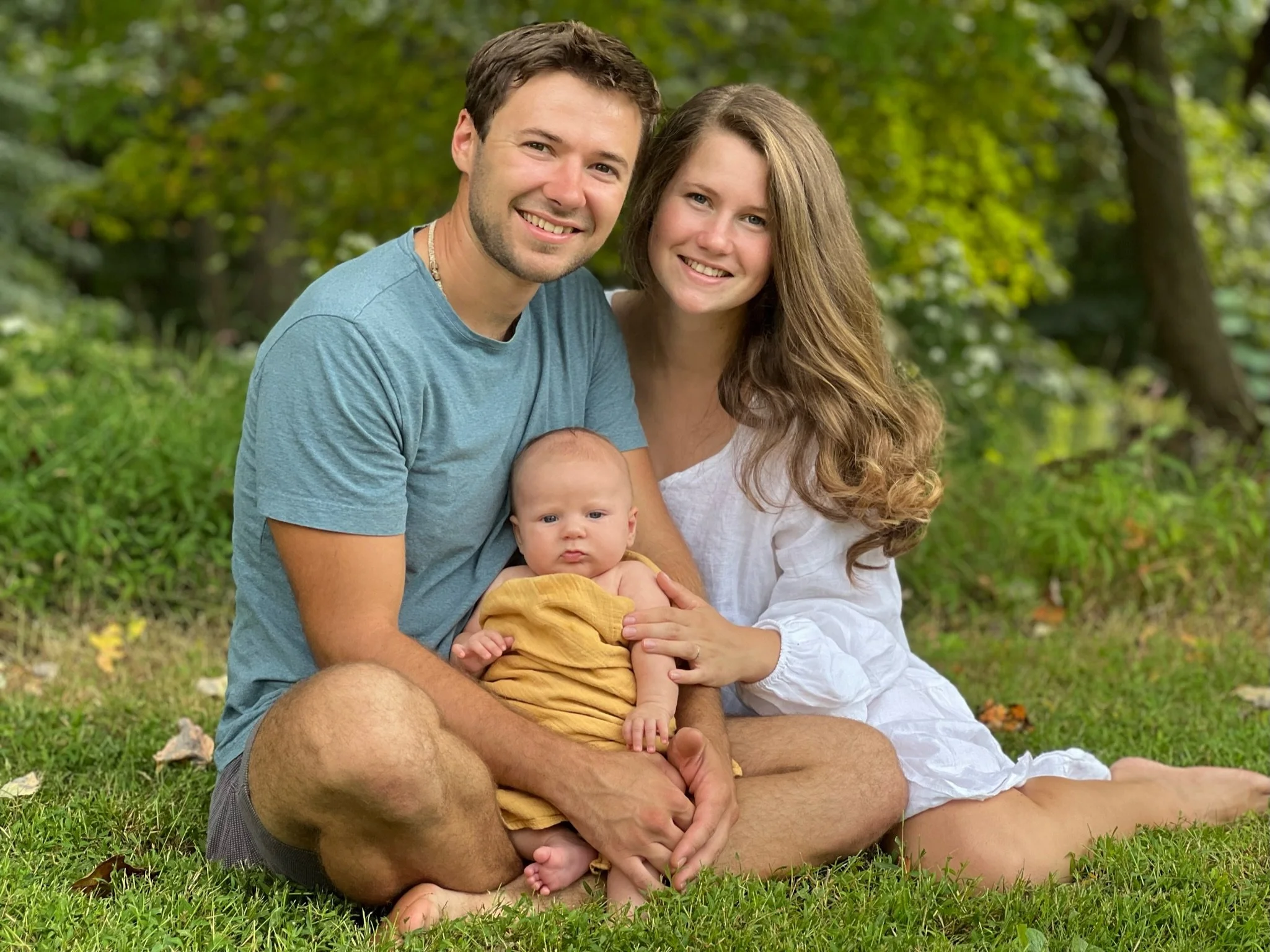 Happy family of three sitting on grass in a park, smiling at the camera, with green trees in the background. The father is wearing a blue t-shirt, the mother a white dress, and their baby is dressed in yellow.