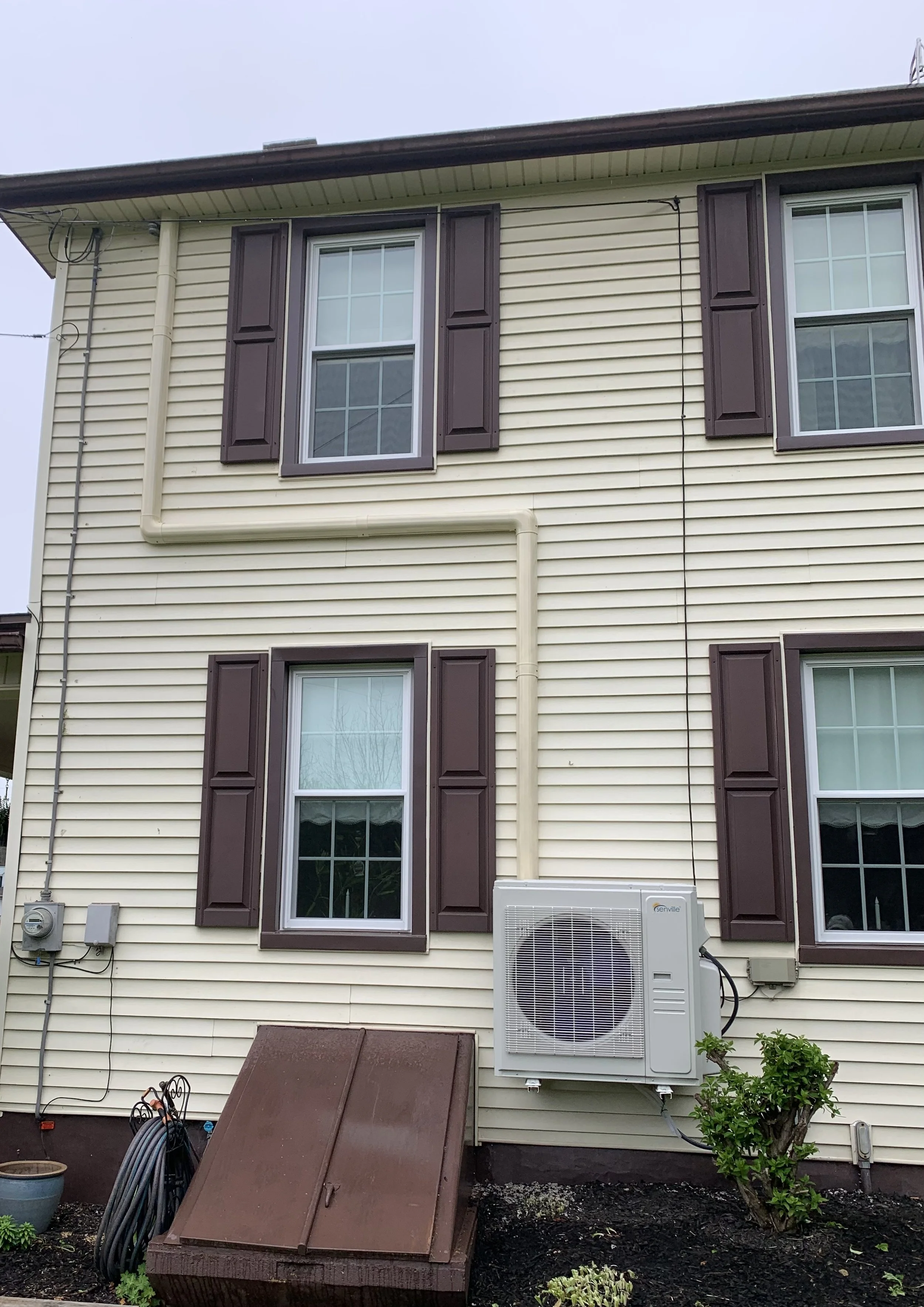 Rear view of a beige, multi-story house with three windows and brown shutters. A white air conditioning unit is attached to the exterior wall near a small green bush. A brown, louvered cover is on the ground, and gardening tools and hose are coiled nearby.