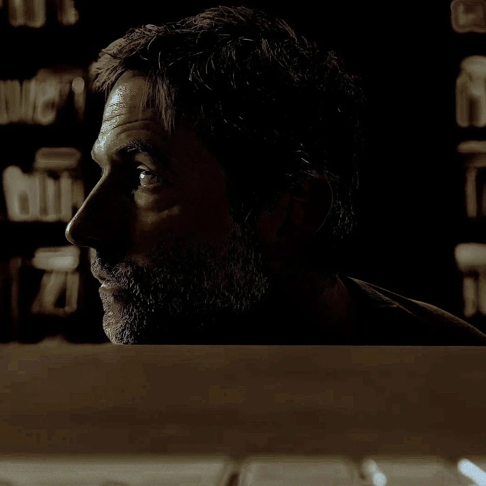 Profile of a man with gray beard and dark hair, sitting in front of a bookshelf, illuminated by soft light in a dark room.