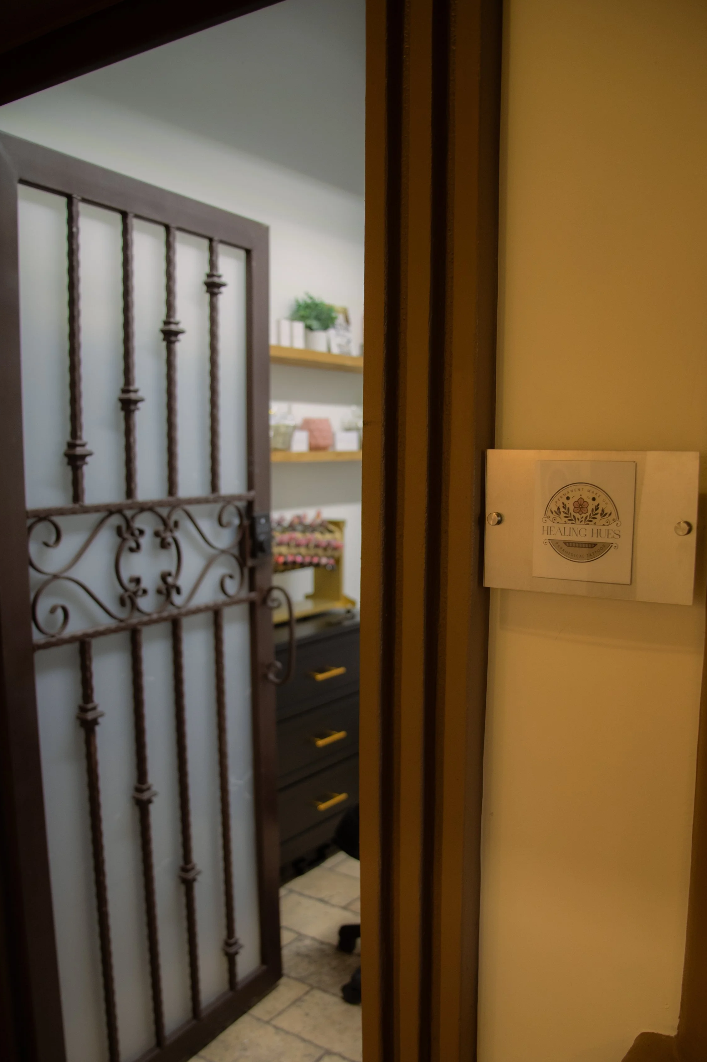 View through an open door into a room with shelves containing neatly organized products and a black cabinet with yellow handles, seen through a metal door with vertical bars.