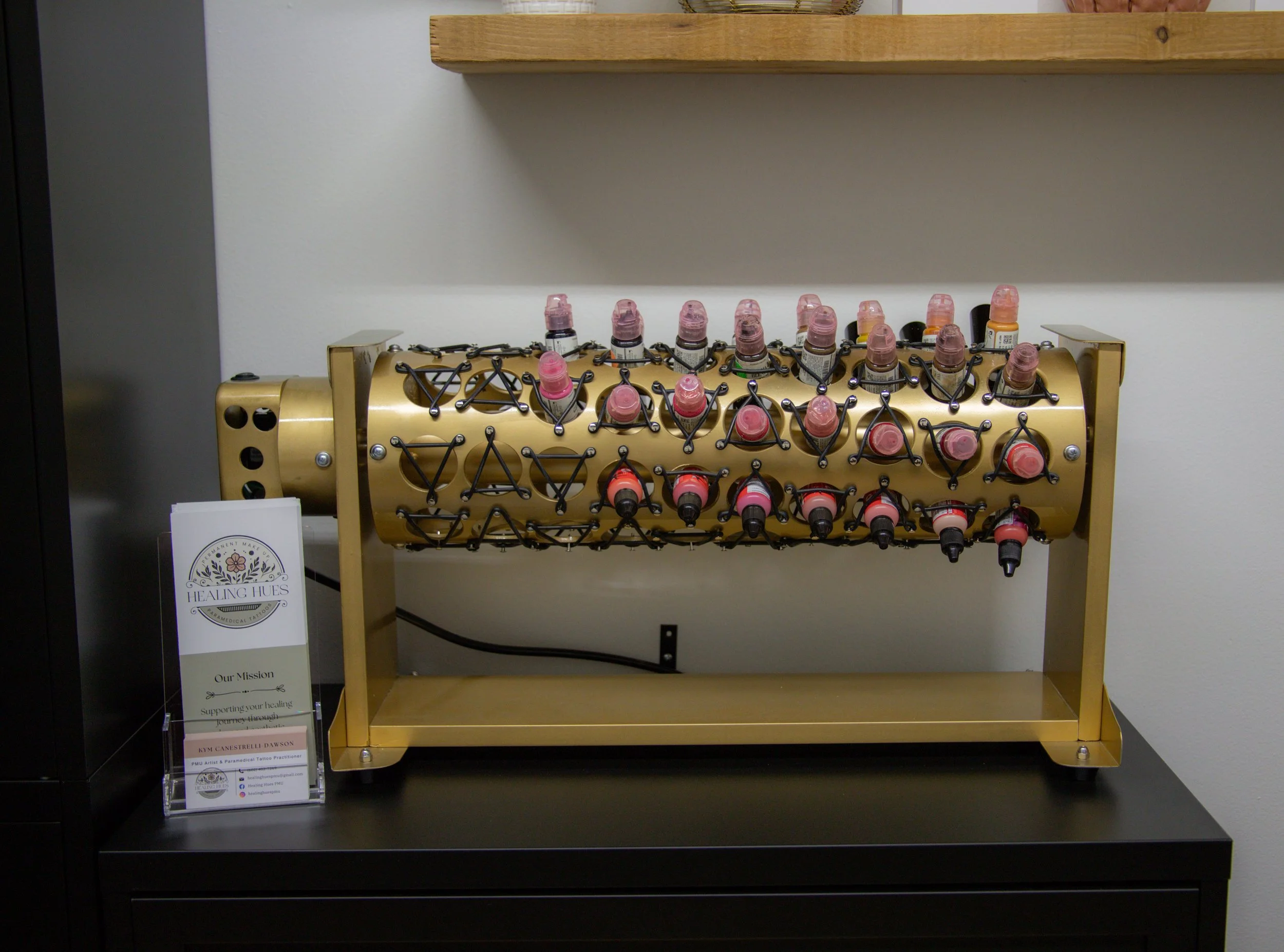 A display of multiple bottles of pink and orange skincare or cosmetic products arranged on a gold-colored cylindrical rack with black metal holders in a retail or clinic setting. A sign next to the display reads 'Healing Hues.'