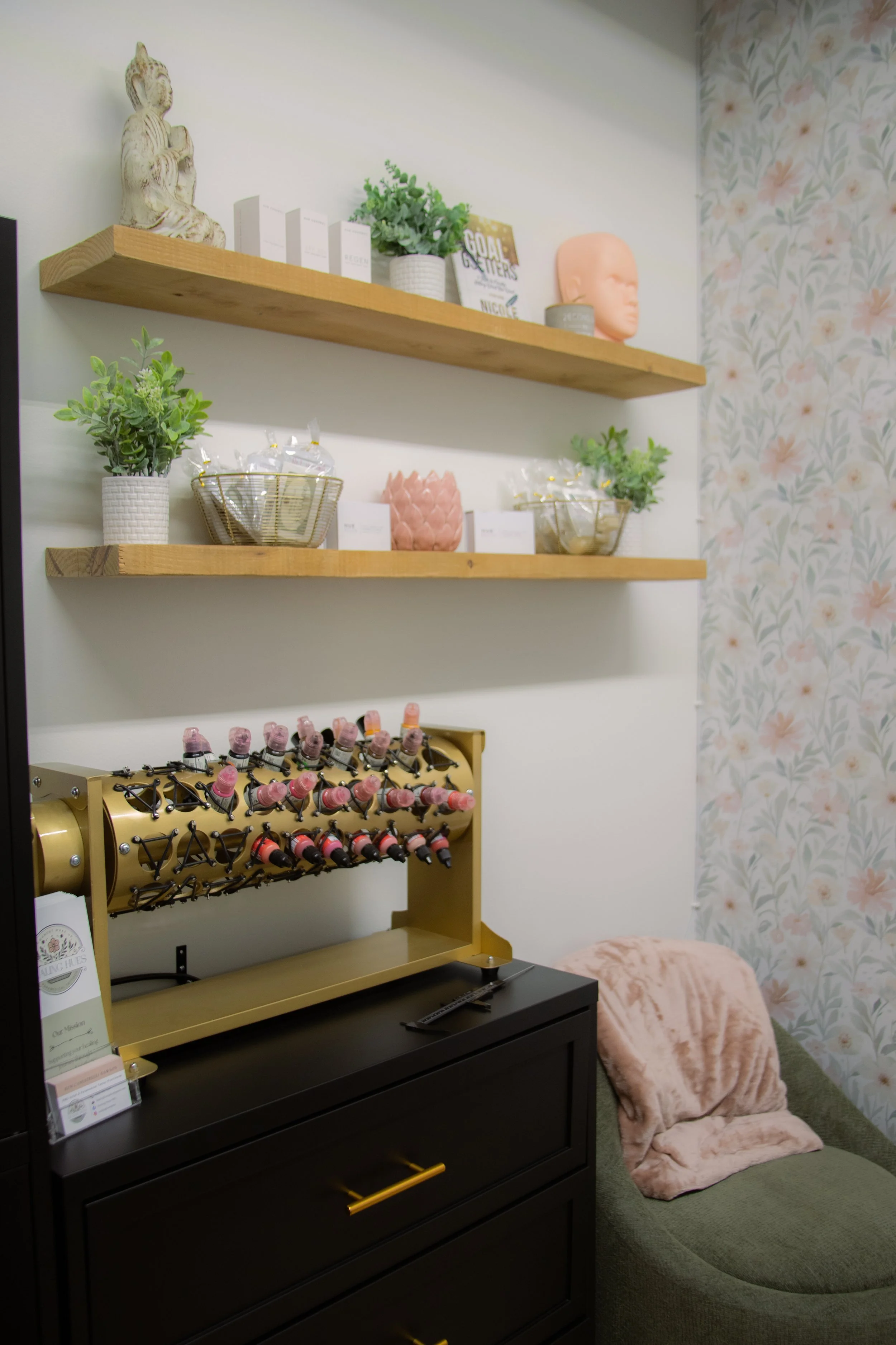 Salon interior featuring two wooden shelves with plants, decorative objects, books, and a mannequin head, a black cabinet with a golden nail polish display, a dark green tufted ottoman, and a pink plush blanket. Floral wallpaper decorates one wall.