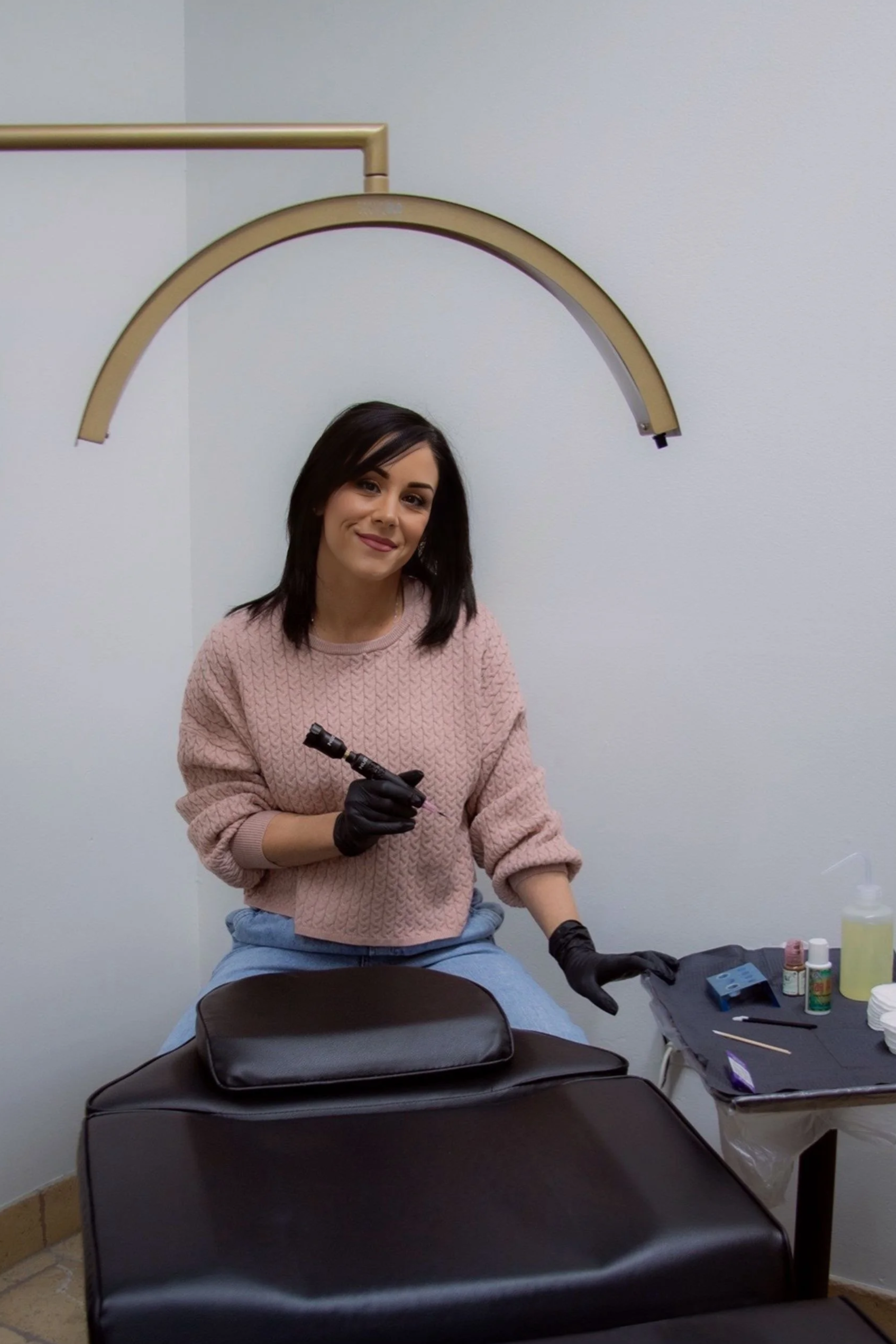 A woman in a pink sweater and gloves sitting at a table with tattooing supplies, smiling, with a large arch overhead and black chairs in the foreground.