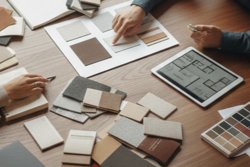 People discussing interior design with fabric and color samples on a wooden table, including a tablet displaying home floor plans.