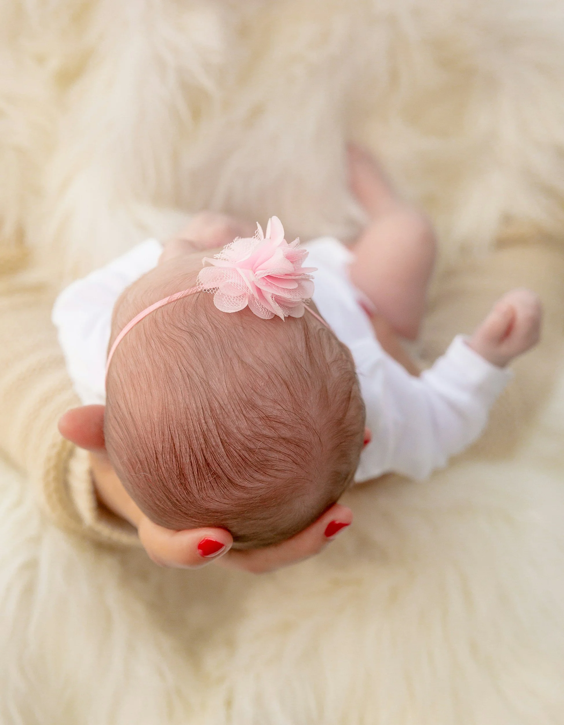 A person holding a newborn baby with a pink flower headband in a white outfit, on a soft, cream-colored surface.