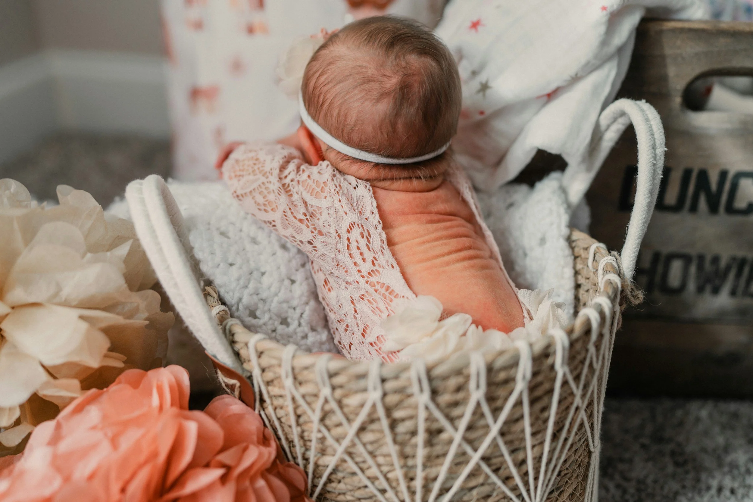A newborn baby girl with a headband sleeping in a small wicker basket, surrounded by soft pink and cream-colored flower decorations.