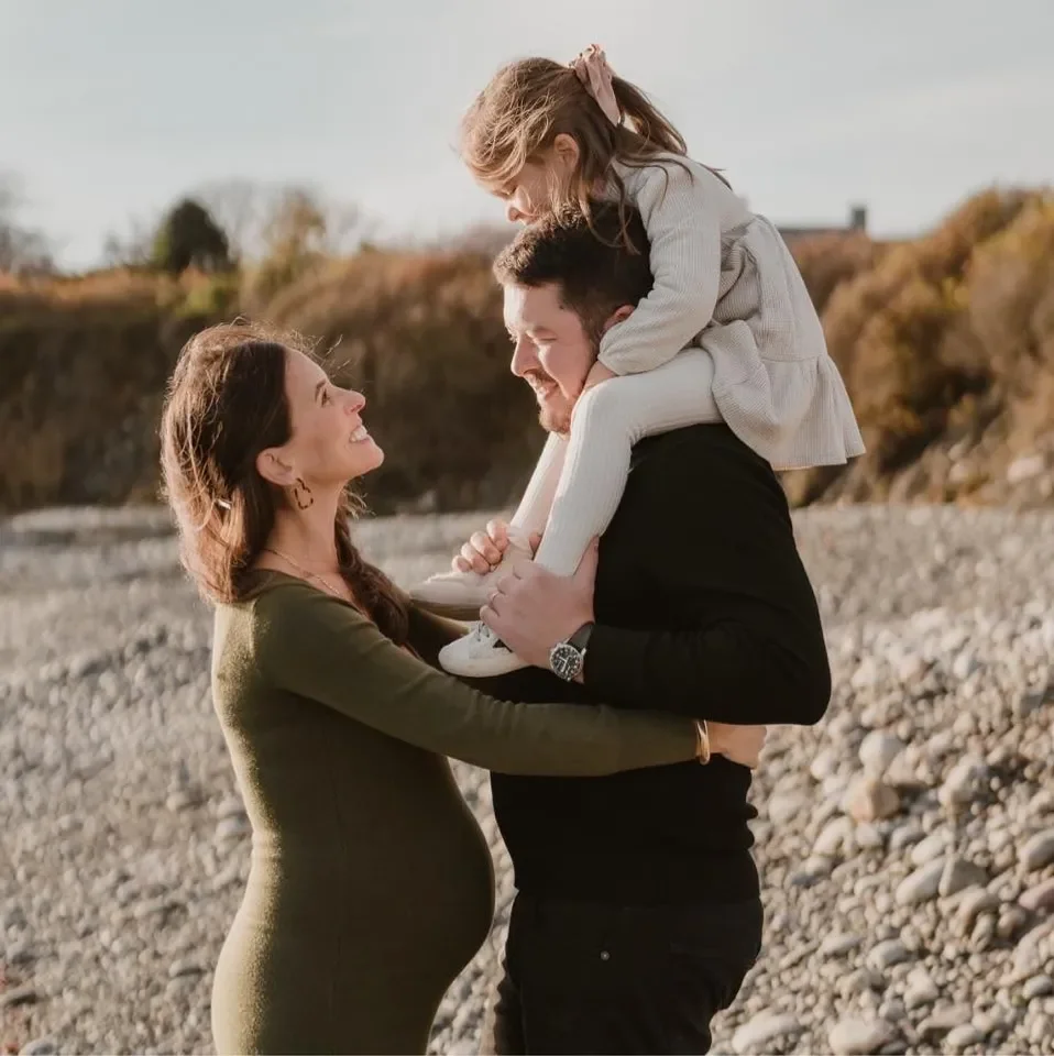 A family of four on a rocky beach, with a pregnant woman smiling at her partner, a man lifting a young girl onto his shoulders, and a young woman looking at her partner.