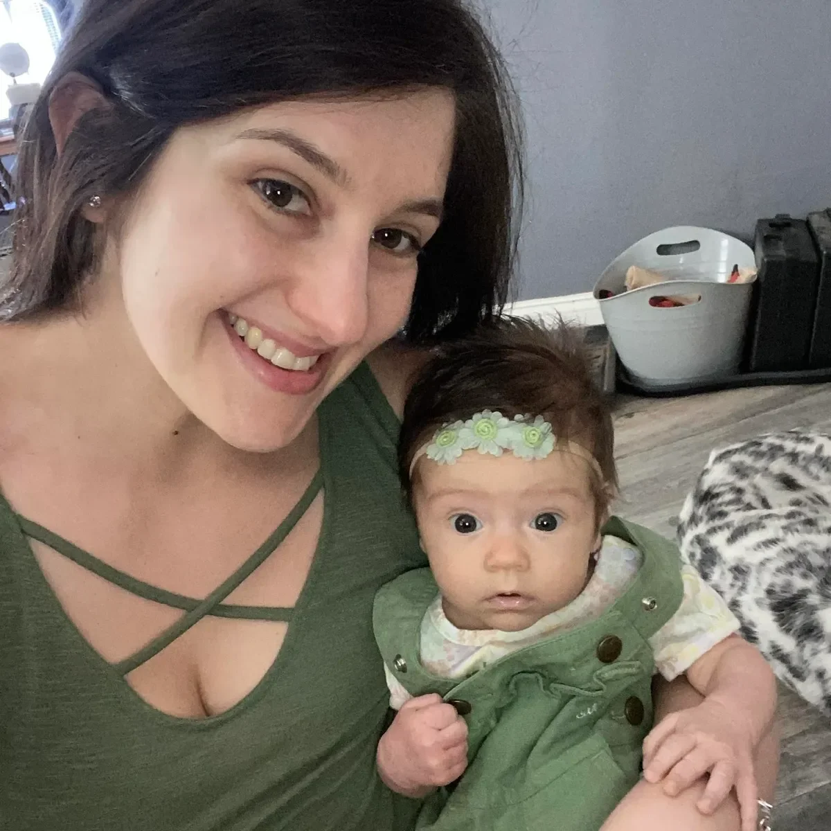 A woman smiling while holding an infant girl with a floral headband and green overalls, in a cozy indoor setting.