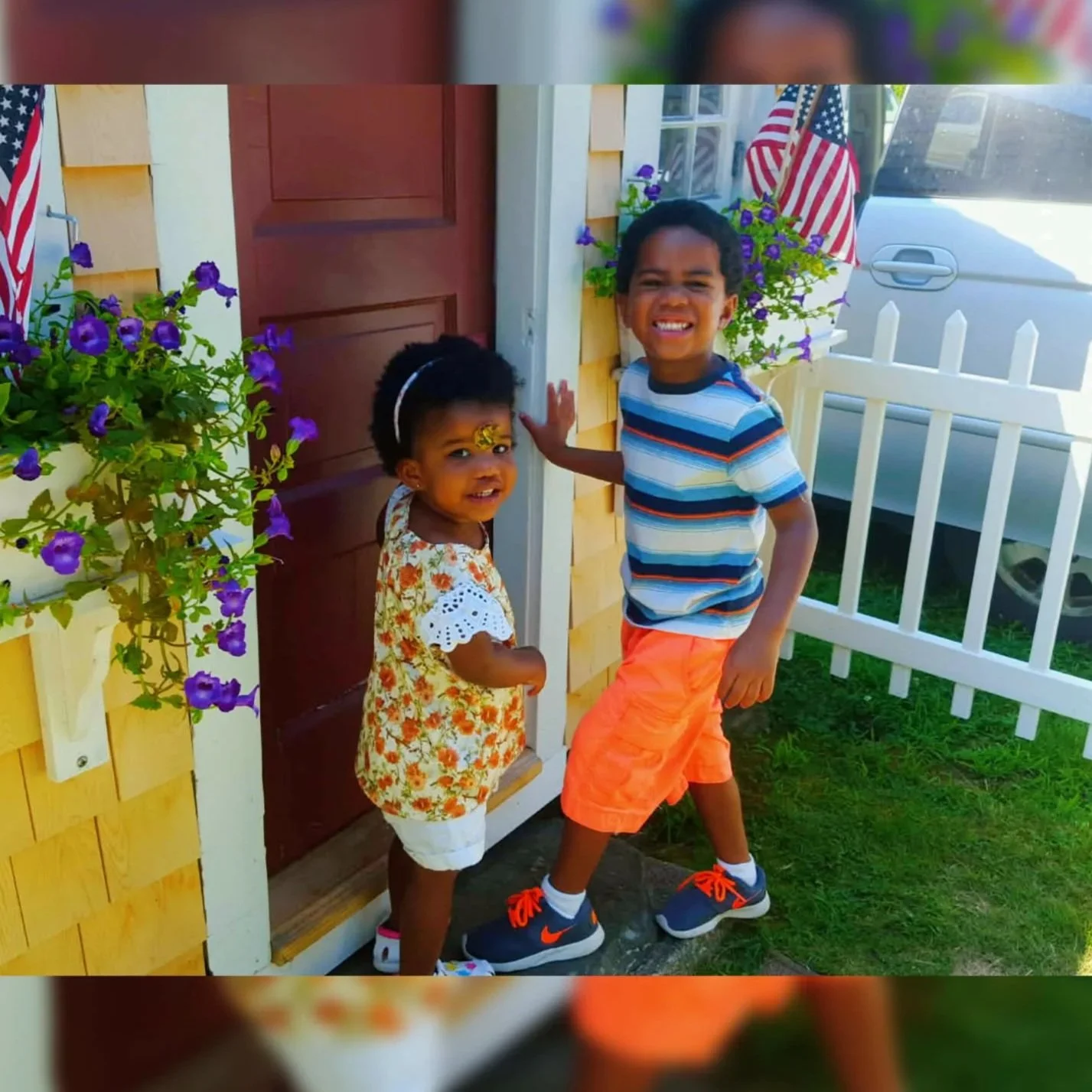 Two children, a girl and a boy, standing at the front door of a house with American flags and purple flowers nearby. The girl is smiling with face paint, and the boy is smiling broadly.