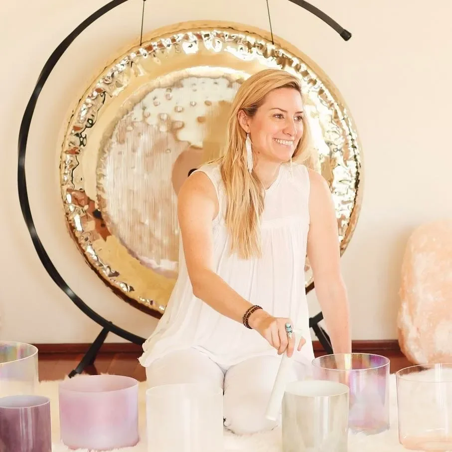 A woman with long blonde hair in a white sleeveless top and white pants, smiling while playing a crystal singing bowl with a mallet. She is sitting on a white surface with several other colored crystal singing bowls around her. Behind her is a large circular mirror with a decorative border, mounted on a stand, reflecting parts of her and the room.
