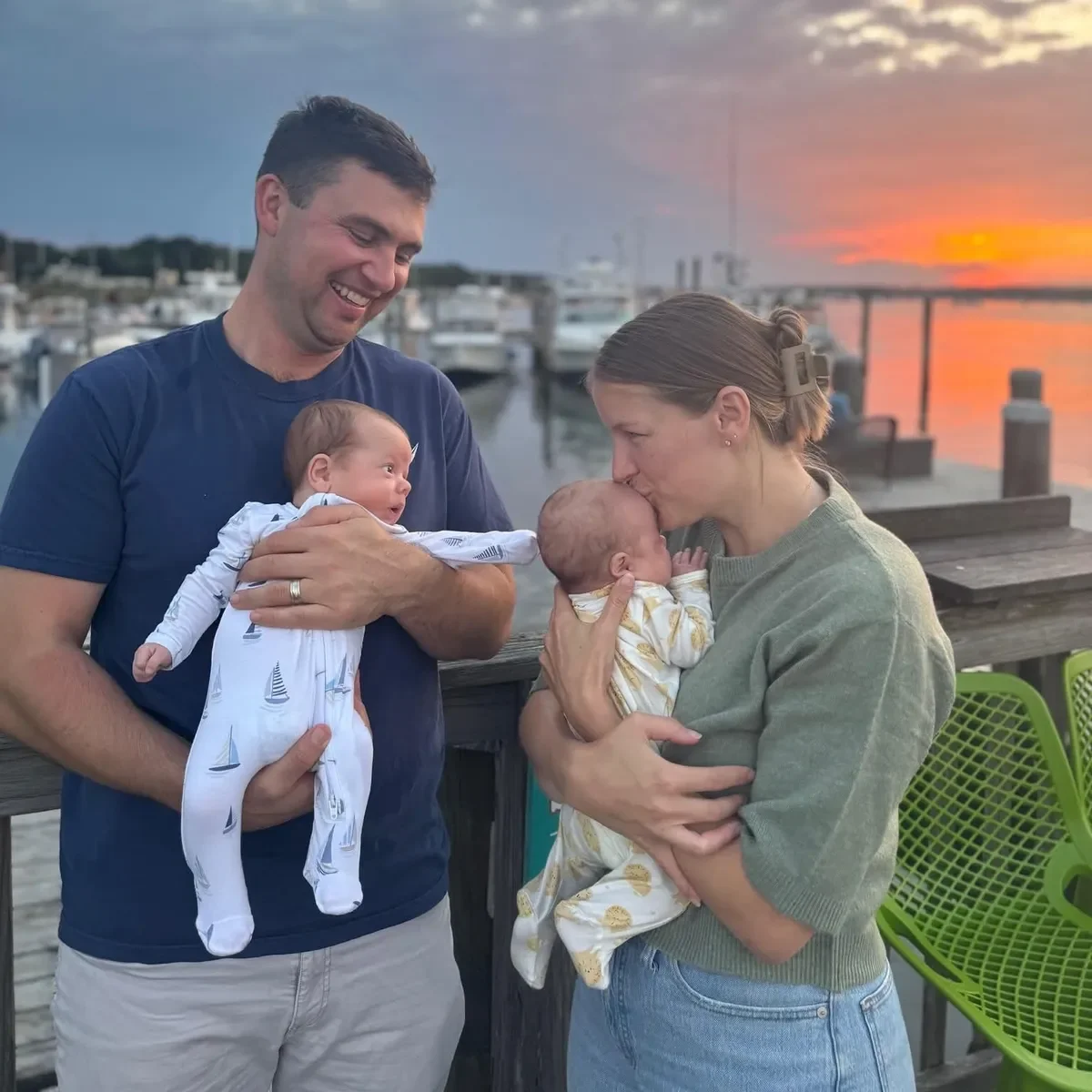 A happy family scene by a marina during sunset, with a man holding a newborn baby, a woman kissing another newborn, and boats docked in the background.