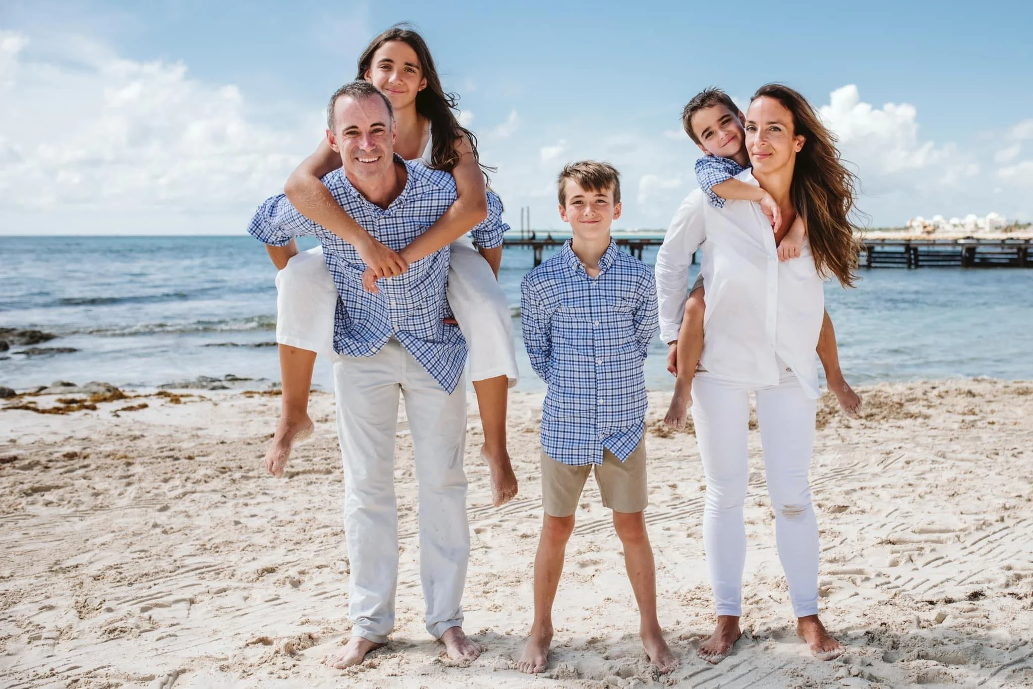A family of five with two parents and three children enjoying a day at the beach, walking on sand with ocean in the background, all smiling and dressed in casual summer clothes.